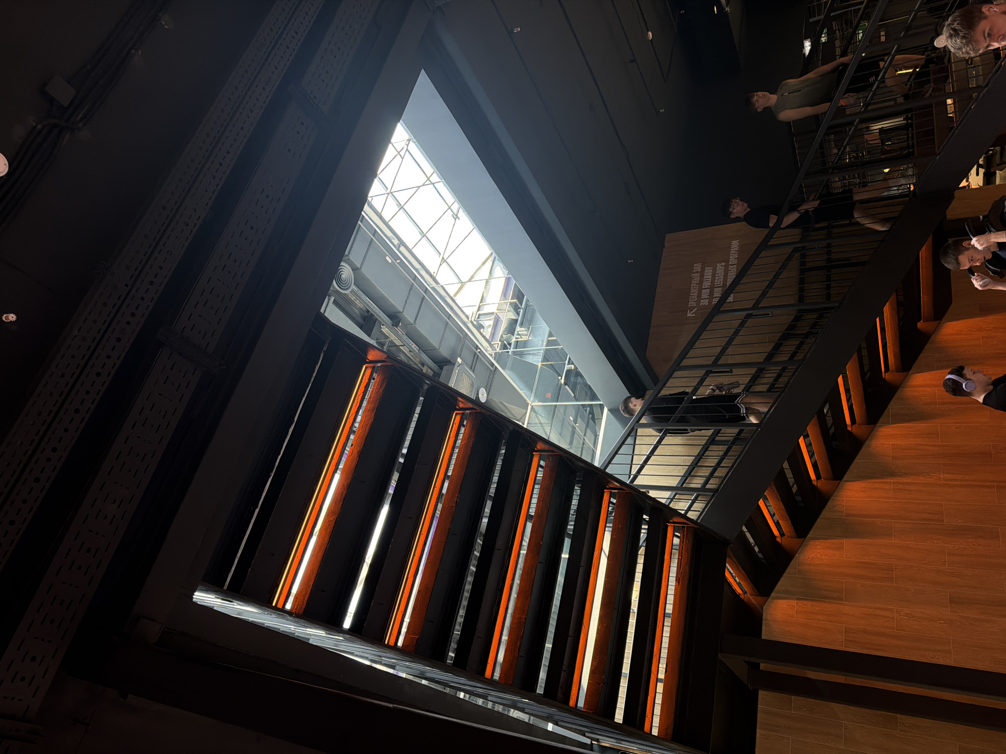 This image captures a dynamic, upward-looking view within a modern, multi-level building, likely a fitness center or gym, characterized by an industrial-chic aesthetic. The perspective is tilted, emphasizing the height and open space.
The main subjects include architectural elements and several people. Dark, perforated metal structures and beams dominate the left and top portions, contrasting with a large, bright skylight at the ceiling, which features a grid pattern and allows ample natural light to flood the space. Below the skylight, a central void is flanked by warm, orange-toned linear lights integrated into dark structural columns, creating striking vertical lines of illumination. On the right, wooden or wood-paneled walls and flooring provide a warm contrast to the dark metalwork.
Several individuals are visible on upper levels, leaning against dark metal railings and observing the space below. One man in a green t-shirt and another in a black t-shirt and glasses are prominent on an upper floor. Further down, another person wearing headphones is partially visible. The people appear relaxed and are not engaged in strenuous activity, suggesting they are either waiting, observing, or moving through the facility.
Notable details include the interplay of natural light from the skylight with artificial orange lighting, casting a distinctive glow on the wooden surfaces. A sign on the wooden wall to the mid-right contains visible Cyrillic text, reading:
- "ПРЕДВАРНЫЙ ЗАЛ" (Preliminary Hall/Room)
- "30 МИН FULLBODY"
- "30 МИН LESSHIPS"
- "БЕДНАЯ ПРОГРАММА" (possibly "Lean Program" or a colloquial phrase in a fitness context, or a playful name, or a misreading of another program name like "Running Program").
This text strongly suggests a fitness-related establishment. The time of day is likely daytime, given the bright natural light. The overall mood is calm and observational. The location context indicates Krasnoyarsk, Russia.