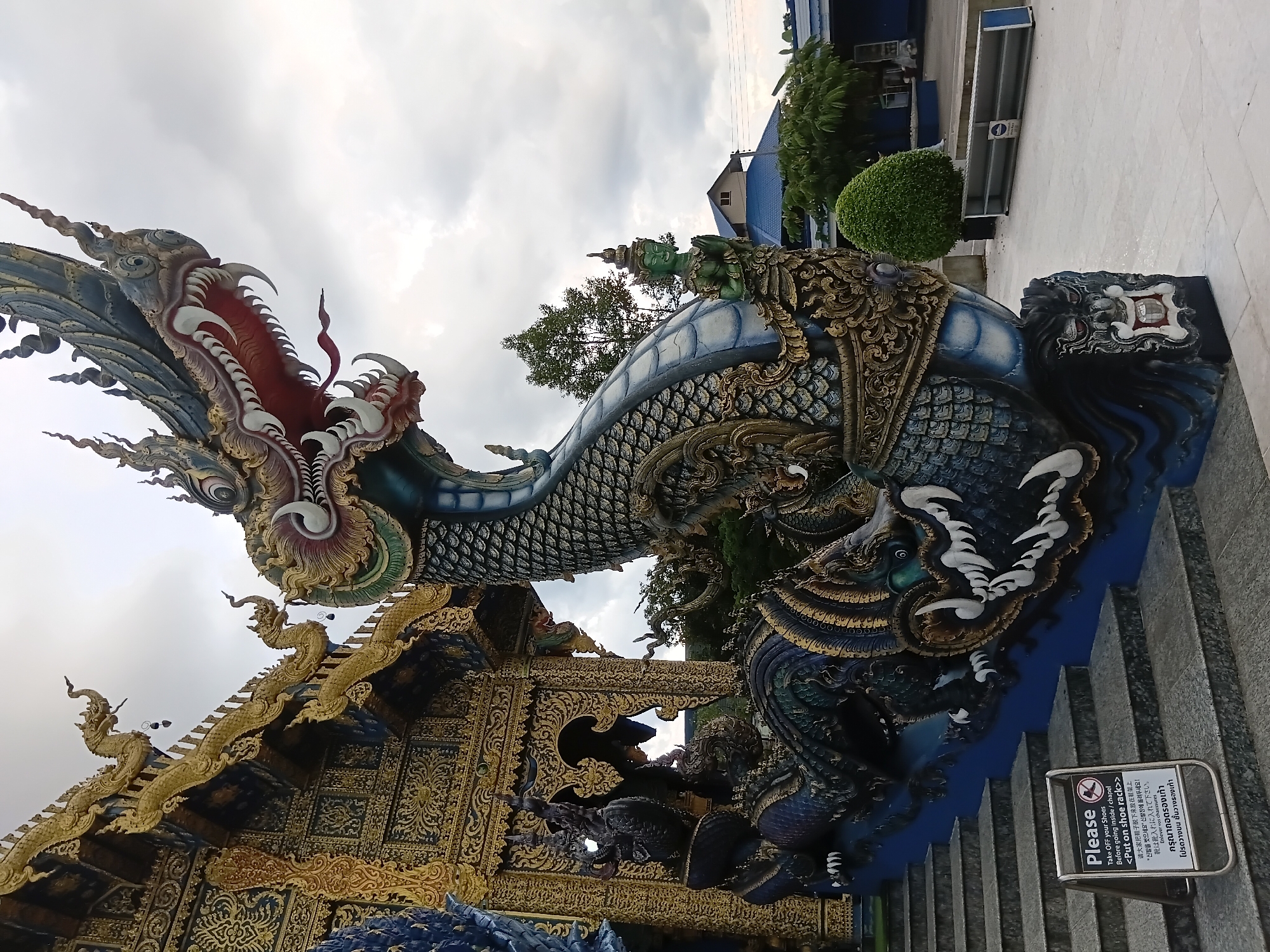 The image depicts a detailed, large-scale sculpture of a mythical Naga serpent, a prominent feature at the entrance to a temple complex in Chiang Rai City Municipality, Thailand. The serpent sculpture, rendered in vibrant blue and gold, coils up a set of stone stairs, its head prominently displayed at the top, with its mouth open revealing sharp teeth and a red tongue. Intricate golden ornamentation adorns the temple's facade in the background, featuring detailed carvings and multiple smaller dragon-like figures along the roofline. A "Please Take Off Your Shoes" sign is visible on the lower steps. To the right, a green-skinned mythical figure is depicted on the serpent's body, with hands clasped in a gesture of greeting. Beyond the temple structures, a paved courtyard, trees, and buildings with blue roofs are visible under a cloudy sky. The overall scene suggests a cultural and religious site, characterized by elaborate artistry and symbolic representations.