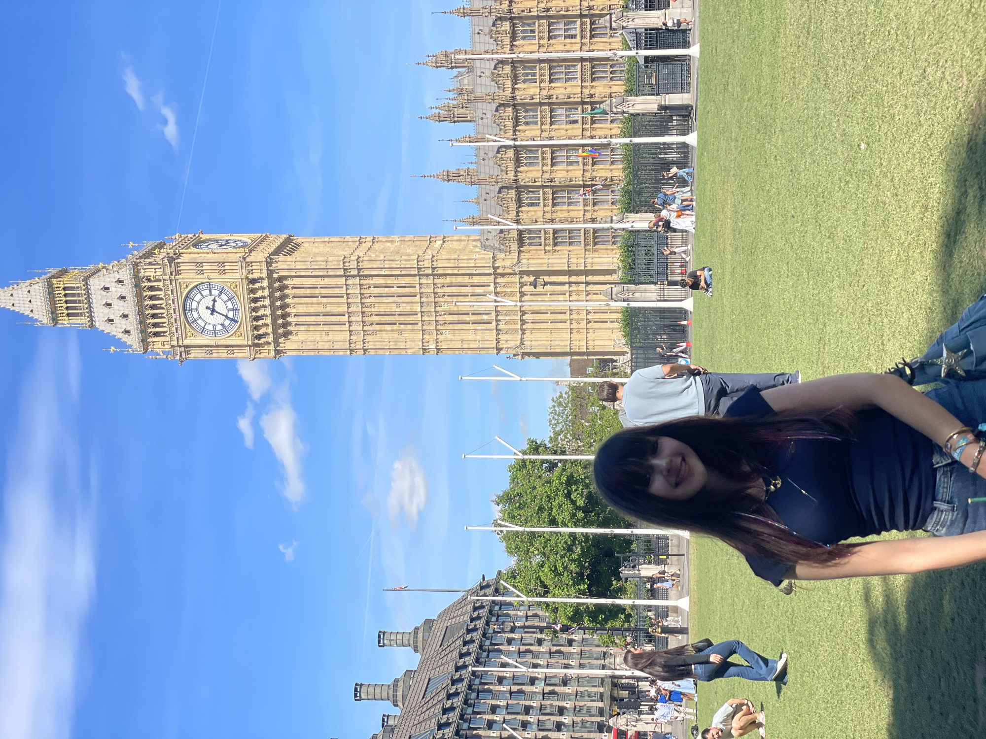 In London, United Kingdom, a young woman with long, dark hair, wearing a navy blue t-shirt and jeans, stands in the foreground of a large grassy area. Behind her, the iconic Elizabeth Tower, commonly known as Big Ben, and the Houses of Parliament dominate the background under a bright blue sky with scattered clouds. Several other people are visible in the midground and background, some walking, others sitting on the grass. The scene is set in an urban park, likely Parliament Square, given the prominent landmarks.