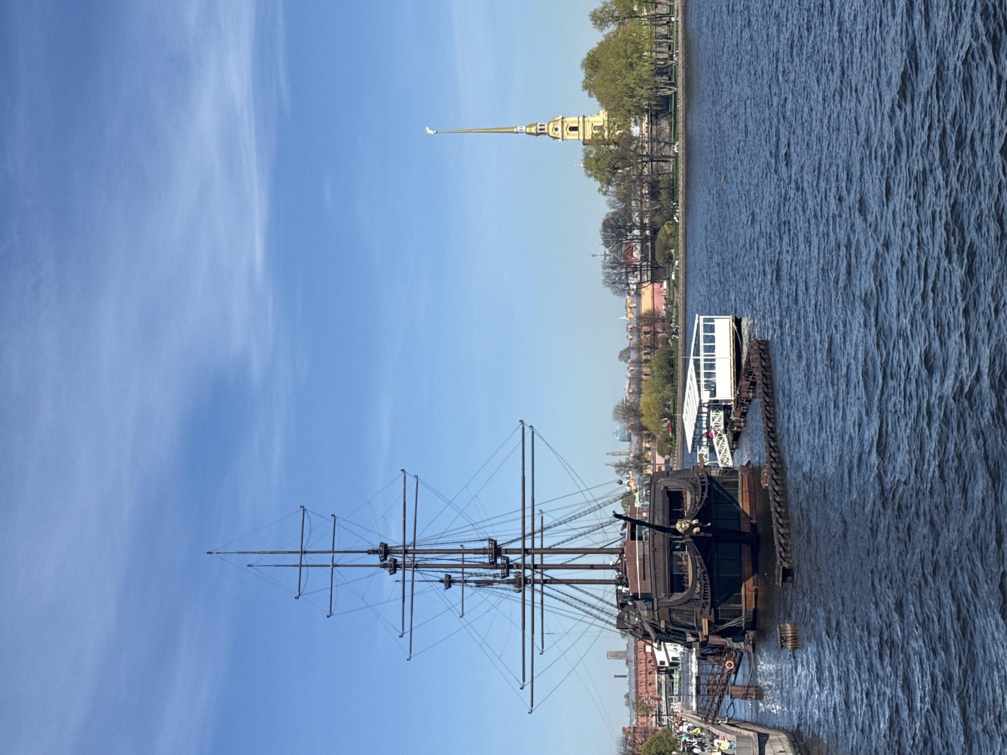 The image shows a tall ship docked on the Neva River in Saint Petersburg, Russia. The Peter and Paul Fortress is visible in the background. The sky is clear blue with some wispy clouds. A white ferry is docked behind the tall ship. People can be seen walking along the embankment.