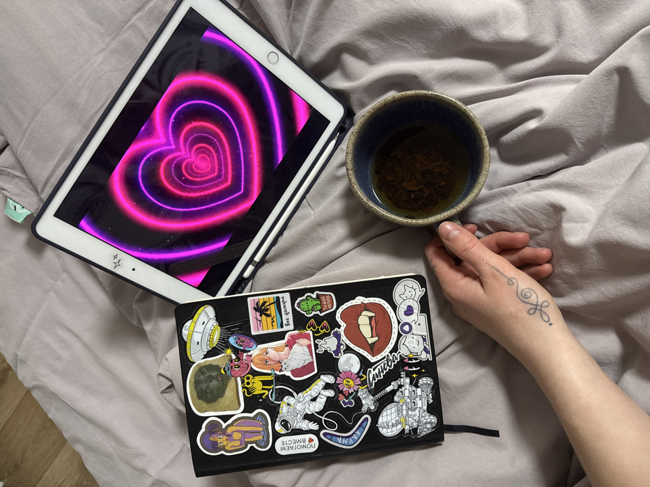 A grey bed features an arrangement of personal items. A white tablet displays a vibrant heart tunnel graphic. A blue ceramic cup filled with dark liquid is next to a hand adorned with a delicate tattoo. A black, sticker-covered notebook rests on the bedspread, adding a casual, artistic touch to the scene.