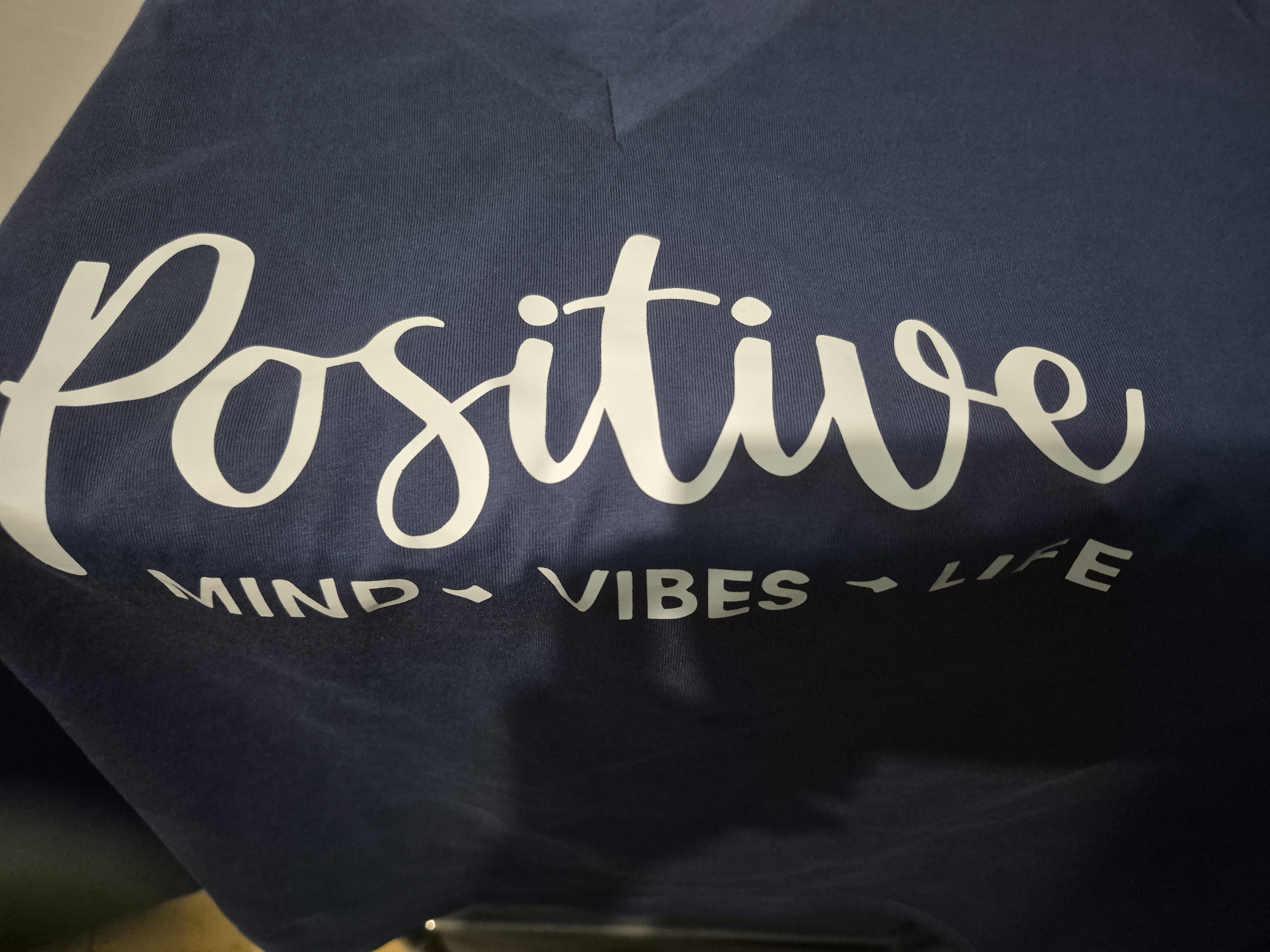 A close-up shot features a dark blue (likely navy) v-neck t-shirt made of a ribbed knit fabric. The shirt has white text printed on it. The main word, "Positive," is written in a large, flowing script font, occupying the central part of the frame. Below "Positive," in a smaller, sans-serif, block letter font, are the words "MIND - VIBES - LIFE," with small arrow-like dashes separating each word. The shirt appears to be indoors, possibly against a plain light-colored wall or background. No individuals or specific activities are visible.