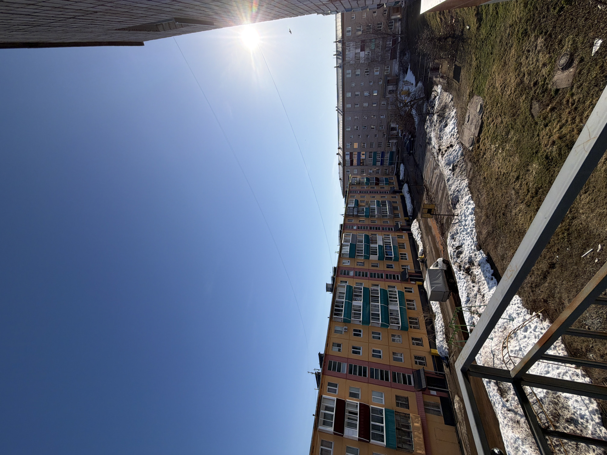 View from a high angle in Sarapul, Russia, capturing a clear blue sky with a bright sun. Below, buildings are visible, including a yellow apartment complex with shuttered windows. Ground view shows patches of snow interspersed with grass and earth. A metal railing is present in the foreground, suggesting the vantage point is from a balcony.