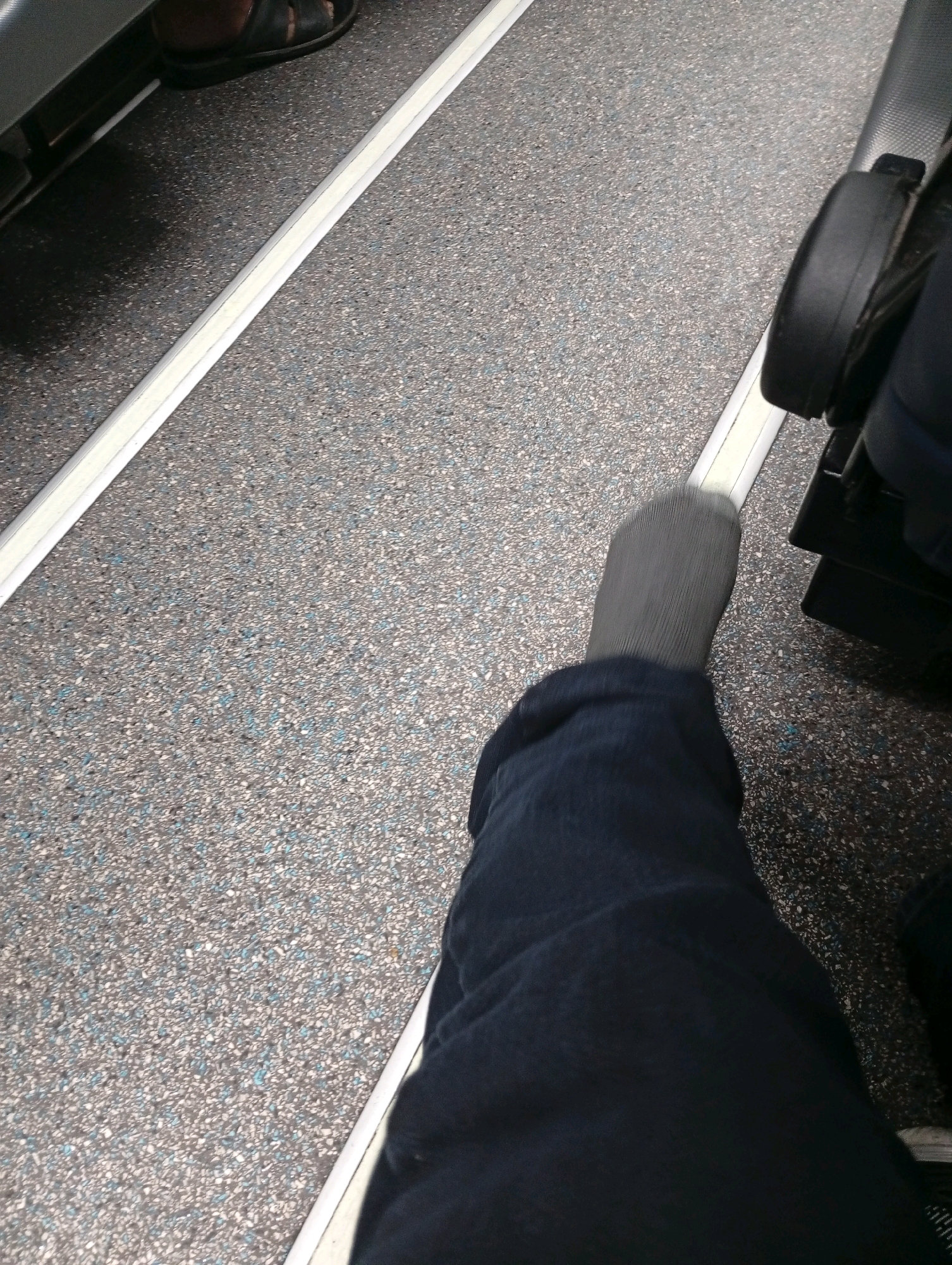 The image shows a close-up view of a person's legs and feet, wearing dark blue pants and grey socks. The person is seated on what appears to be a form of public transportation, possibly a train or bus, as indicated by the textured flooring and the presence of a black seat structure to the right. A white strip runs along the floor, separating the seating area. The environment is dimly lit, with the focus on the lower half of the body. The image does not contain any specific landmarks or details that would allow for identification of its location within Silihasih, Indonesia.