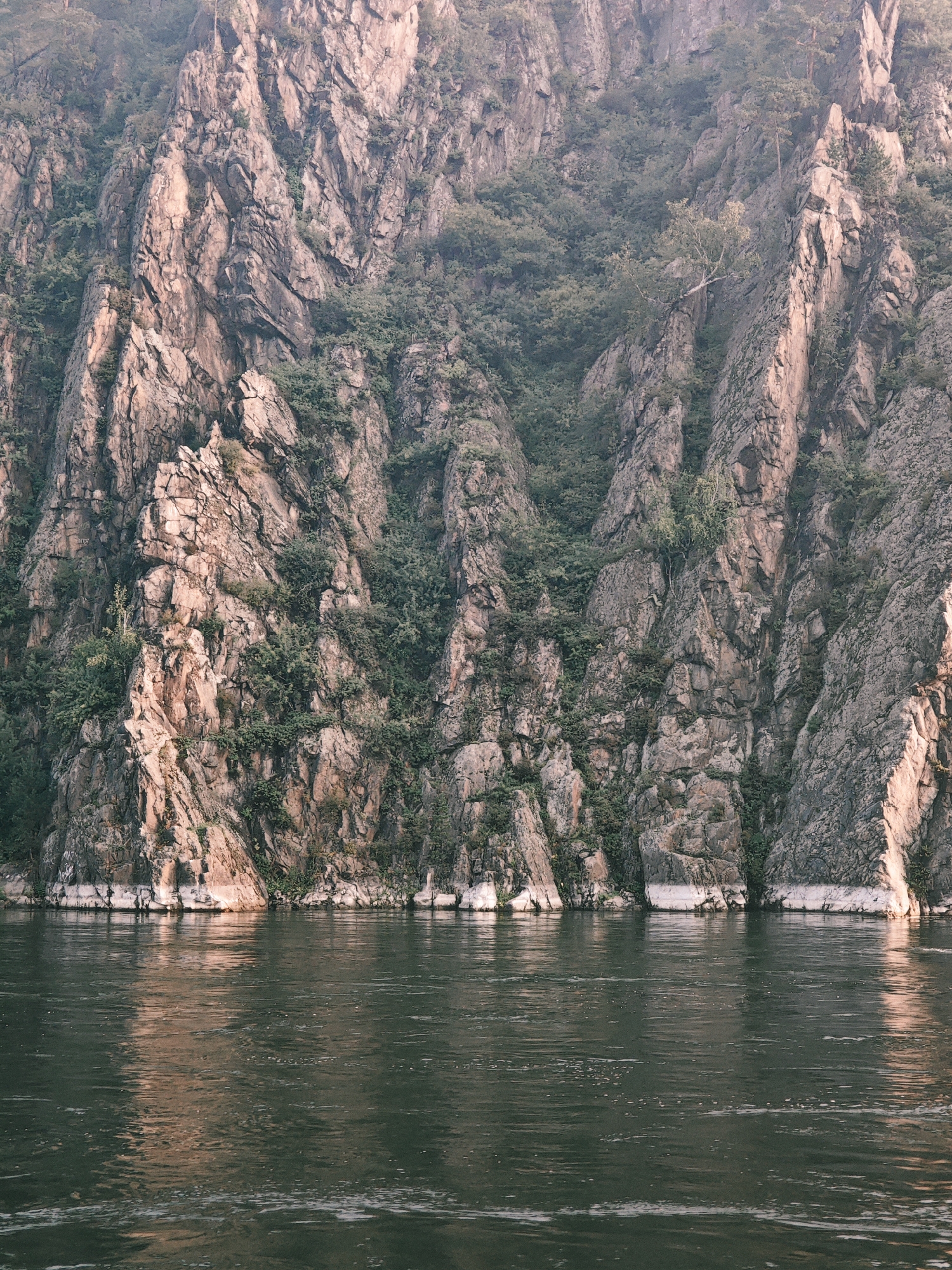 A wide, dark river flows in the foreground, reflecting the muted light of the sky. Behind the river, a steep, rocky cliff rises dramatically, covered in patches of dense green foliage and sparse trees. The rock face is textured and weathered, with visible cracks and crevices. The scene depicts a natural landscape along the riverbank in or near Atamanovo, Russia.