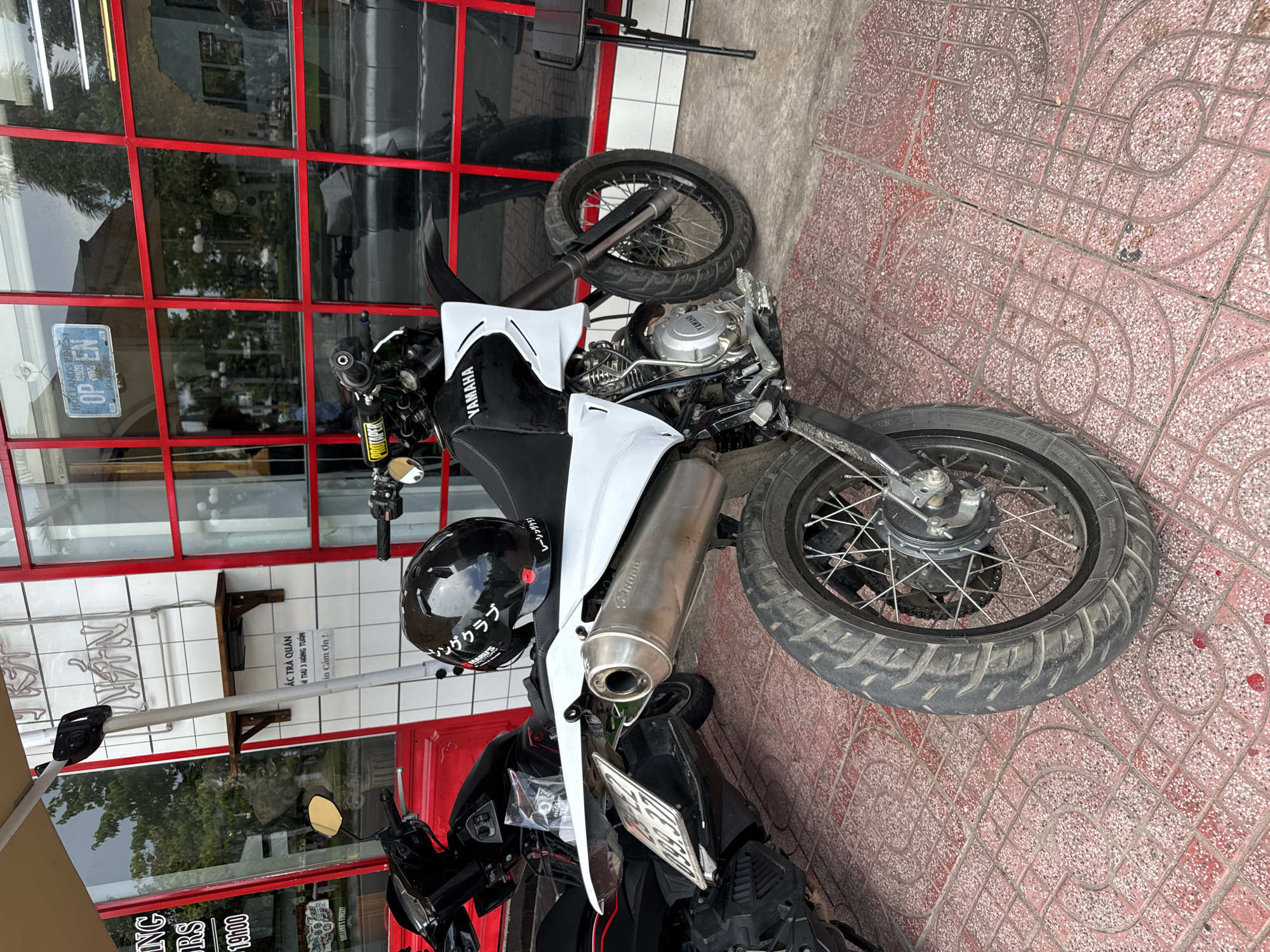 In Phan Thiết, Vietnam, a white and black dirt bike is parked on a red tiled sidewalk in front of a building with red-framed windows. The bike has a silver exhaust pipe, a black helmet with white lettering on the seat, and a license plate that is partially visible. Behind the bike, a black scooter is parked. The building appears to be a shop or restaurant, with signs in Vietnamese on the wall. A black metal chair is visible to the right of the bike.