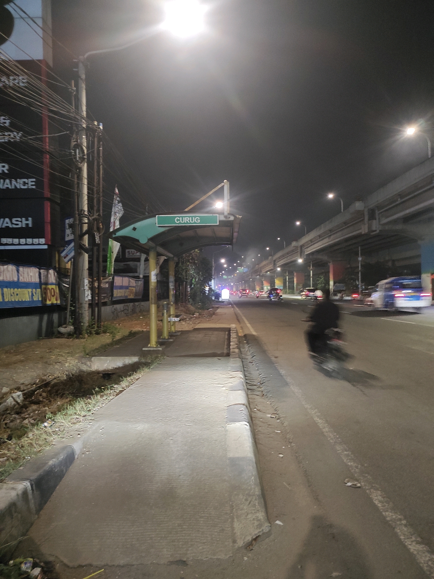 An outdoor, nighttime shot captures a street scene in East Jakarta, Indonesia, likely near a bus stop. The foreground is dominated by a concrete sidewalk and a dirt embankment. A bus stop shelter with the word "CURUG" visible on its sign stands to the right, supported by yellow poles. Beyond the sidewalk, a busy road is visible, filled with moving vehicles, including cars and a motorcycle that appears blurred due to motion. An elevated highway structure looms in the background, illuminated by streetlights and the headlights of passing vehicles. The overall atmosphere is urban and active, with the darkness of night punctuated by artificial light sources.