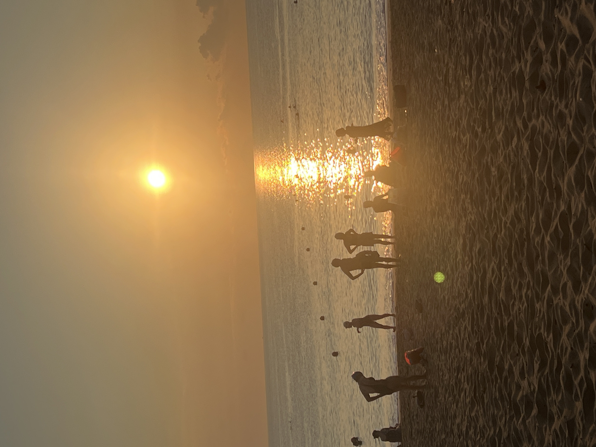 The image captures a vibrant sunset scene on a sandy beach, likely in a coastal location like Nha Trang, Vietnam, given the golden hour light and tropical feel. The sun, a bright yellow orb, dominates the upper left of the frame, casting a brilliant, shimmering path of light across the calm water of the sea. The sky above is a gradient of warm oranges and yellows, with a few faint clouds visible in the upper right.
Numerous people, mostly adults and some children, are silhouetted against the bright sun and reflective water. They are scattered along the shoreline, either standing in the shallow water or on the wet sand close to the water's edge. Their forms are dark and indistinguishable, but they appear to be enjoying the evening, some seemingly wading or strolling.
The foreground features the textured, undulating patterns of wet sand, indicating the receding tide. Several small, dark buoys are visible floating in the water, extending into the distance. A reddish object, possibly a bag or toy, is partially visible on the sand near one of the silhouetted figures. Another curious detail is a bright green, circular glow on the sand in the mid-ground, which could be a light source or a lens flare.
The overall atmosphere is warm, peaceful, and leisurely, characteristic of a tropical beach at dusk. No visible text is present in the image.