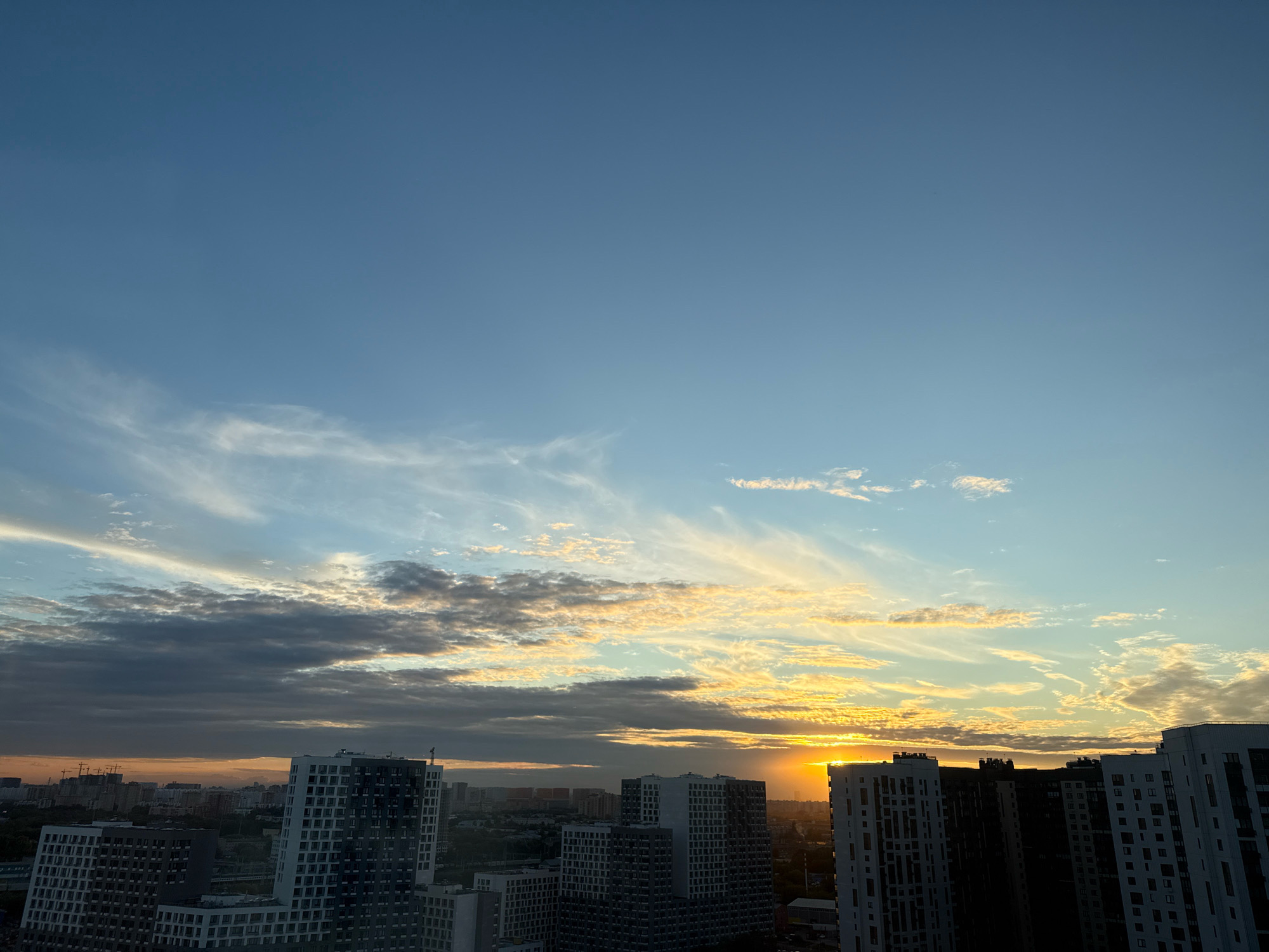The image captures a sunset over the city of Lyubertsy, Russia. The sky is a gradient of blues, transitioning to warm oranges and yellows as the sun dips below the horizon. Wispy clouds are illuminated by the setting sun, creating streaks of light across the sky. In the foreground, several modern apartment buildings with geometric designs rise above the city skyline. The buildings are illuminated by the ambient light of the sunset. The lower portion of the image shows the cityscape of Lyubertsy, with more buildings and some construction cranes visible in the distance. The overall impression is one of an urban environment bathed in the dramatic light of a sunset.