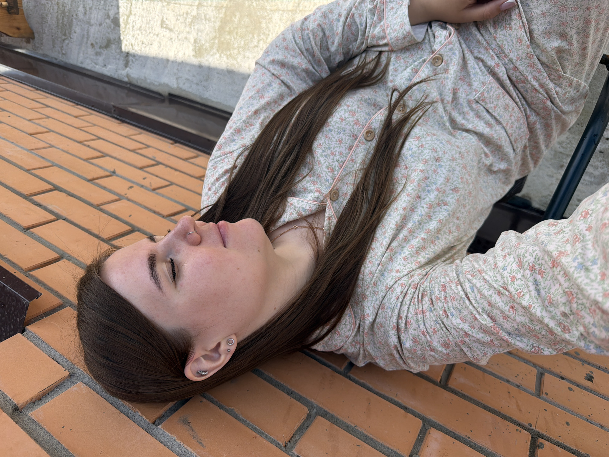 A young woman with long brown hair is lying down on a brick surface, her head tilted back. She is wearing a light-colored pajama top with a floral print featuring small pink and blue flowers. Her eyes are closed, and her expression appears relaxed or peaceful. She has a nose ring and multiple earrings in her left ear. The background shows a textured wall and a metal gutter, suggesting an outdoor setting, possibly a rooftop or balcony. The lighting suggests it is daytime, with sunlight casting shadows on the brickwork. The scene is static, with no discernible activity other than the woman resting. The location is identified as Chaikovsky, Russia. There is no visible text in the image.
