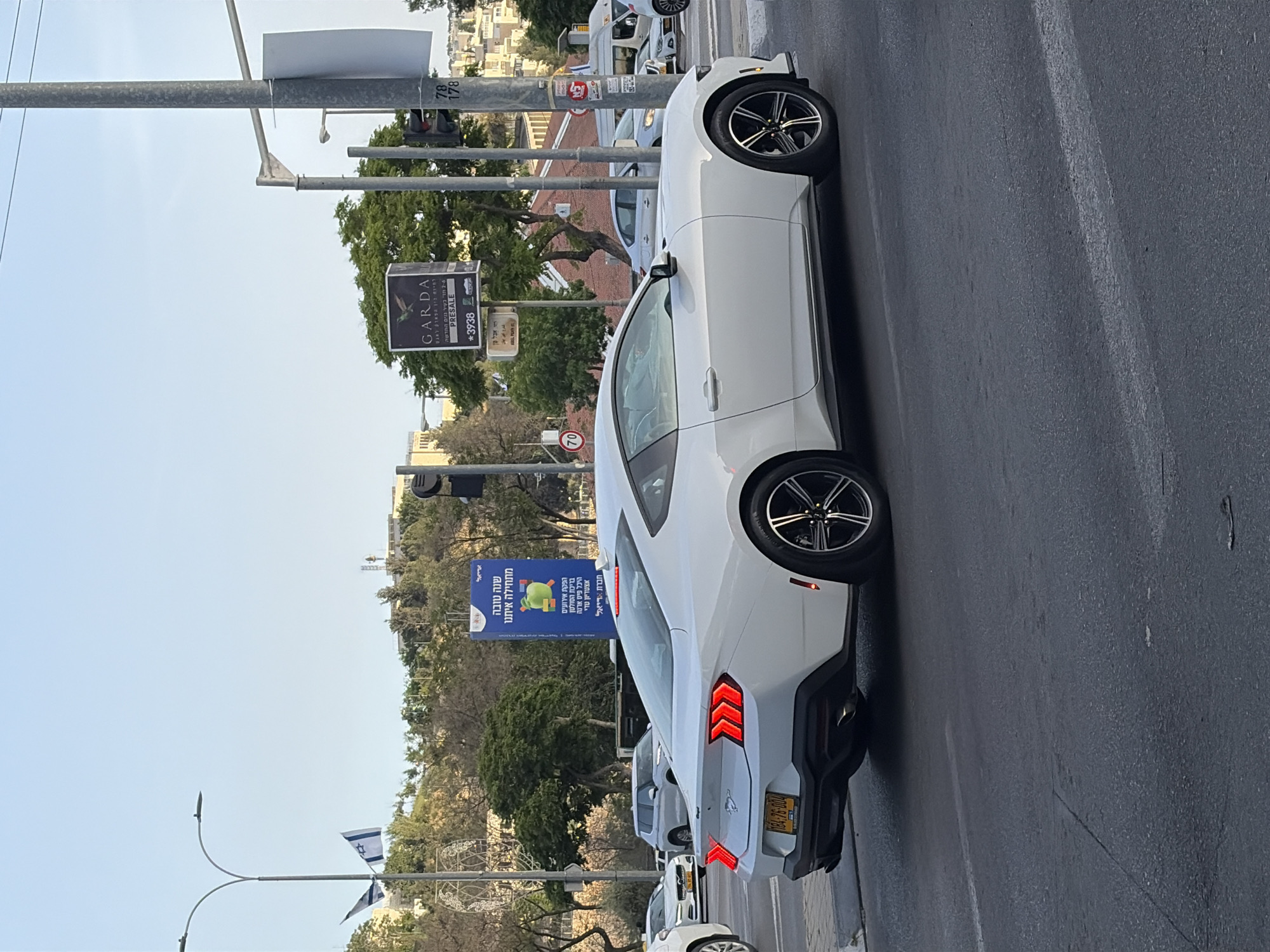 The image shows a white car, likely a Ford Mustang, parked on the side of a road. The license plate is visible: 84-76-004. Roadside signage in Hebrew can be seen in the background. A Garda Pre-Sale sign is visible. There is a building sign in Hebrew, as well. An Israeli flag can be seen by a decorative street light. There is traffic visible in the image. The image appears to be taken in the city of Jerusalem, Israel.