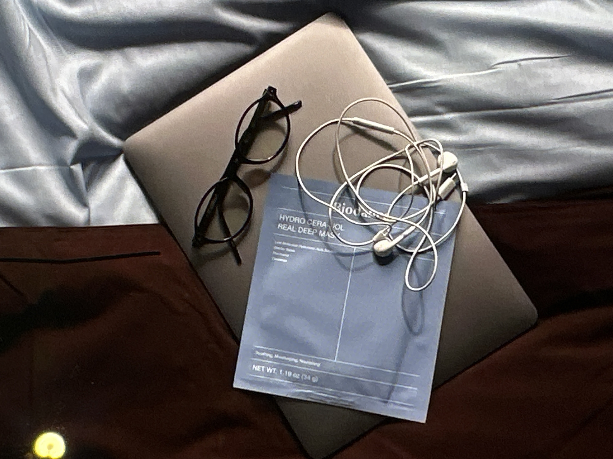 The image shows a gray laptop on a bed with a light blue sheet and brown blanket. On top of the laptop, there is a pair of black eyeglasses, a set of tangled white earbuds, and a blue Biologique Recherche sheet mask labeled "Hydro Cera-Iol Real Deep Mask". The net weight of the mask is listed as 1.19 oz (34 g). The environment appears to be indoors and domestic. The location is not deducible from the given information.