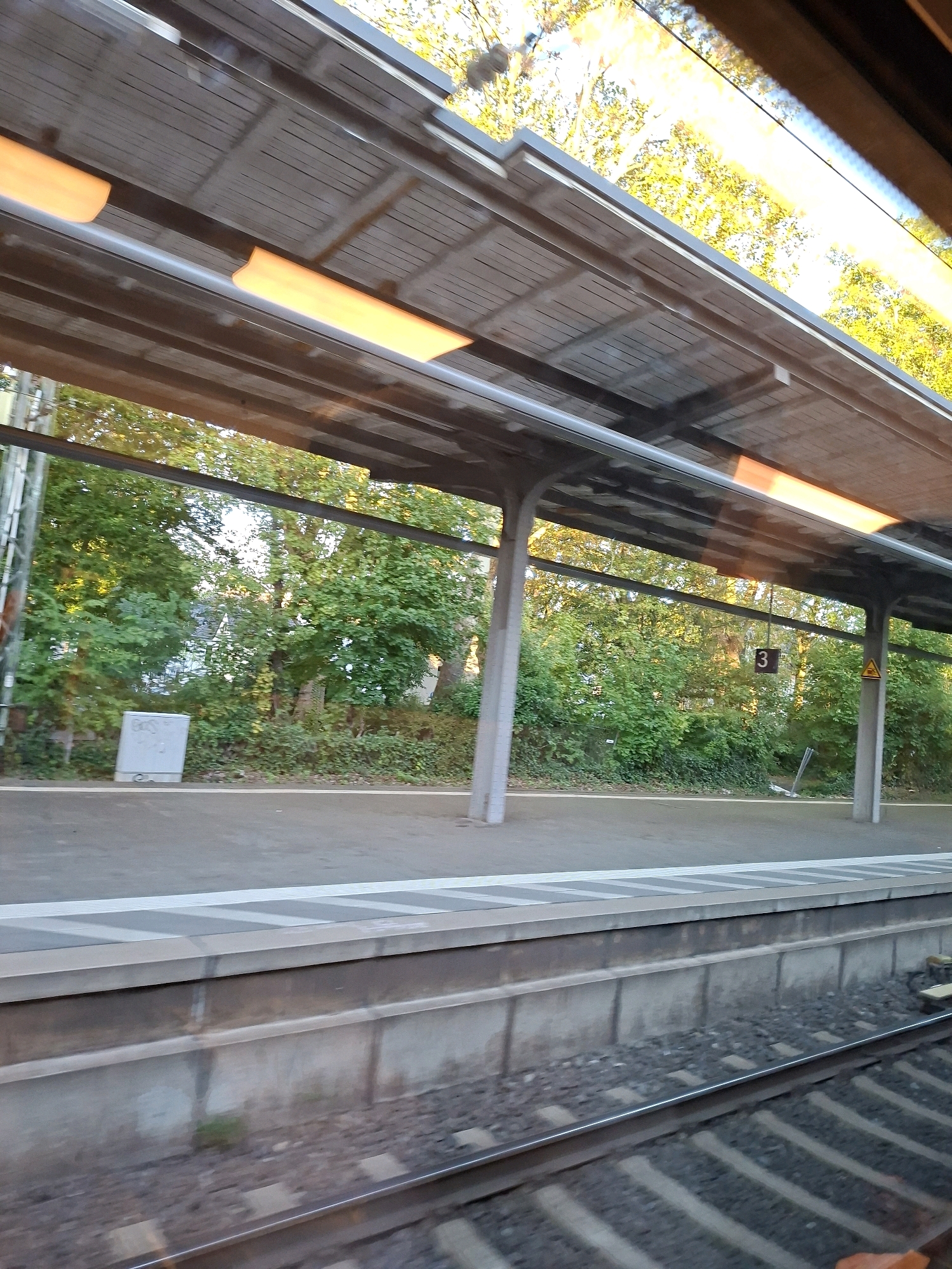 This image captures a view of a train station platform in Bonn, Germany, looking towards lush green trees and buildings in the background. The platform is covered by a modern canopy with visible fluorescent lighting. A light gray utility box is positioned on the platform to the left, near the trees. A sign with the number "3" is hanging from the canopy, along with a yellow triangular warning sign. The train tracks are visible in the foreground, with ballast and ties. The overall scene suggests an outdoor setting during daylight hours, possibly late afternoon given the warm lighting. There are no people visible in the image.
