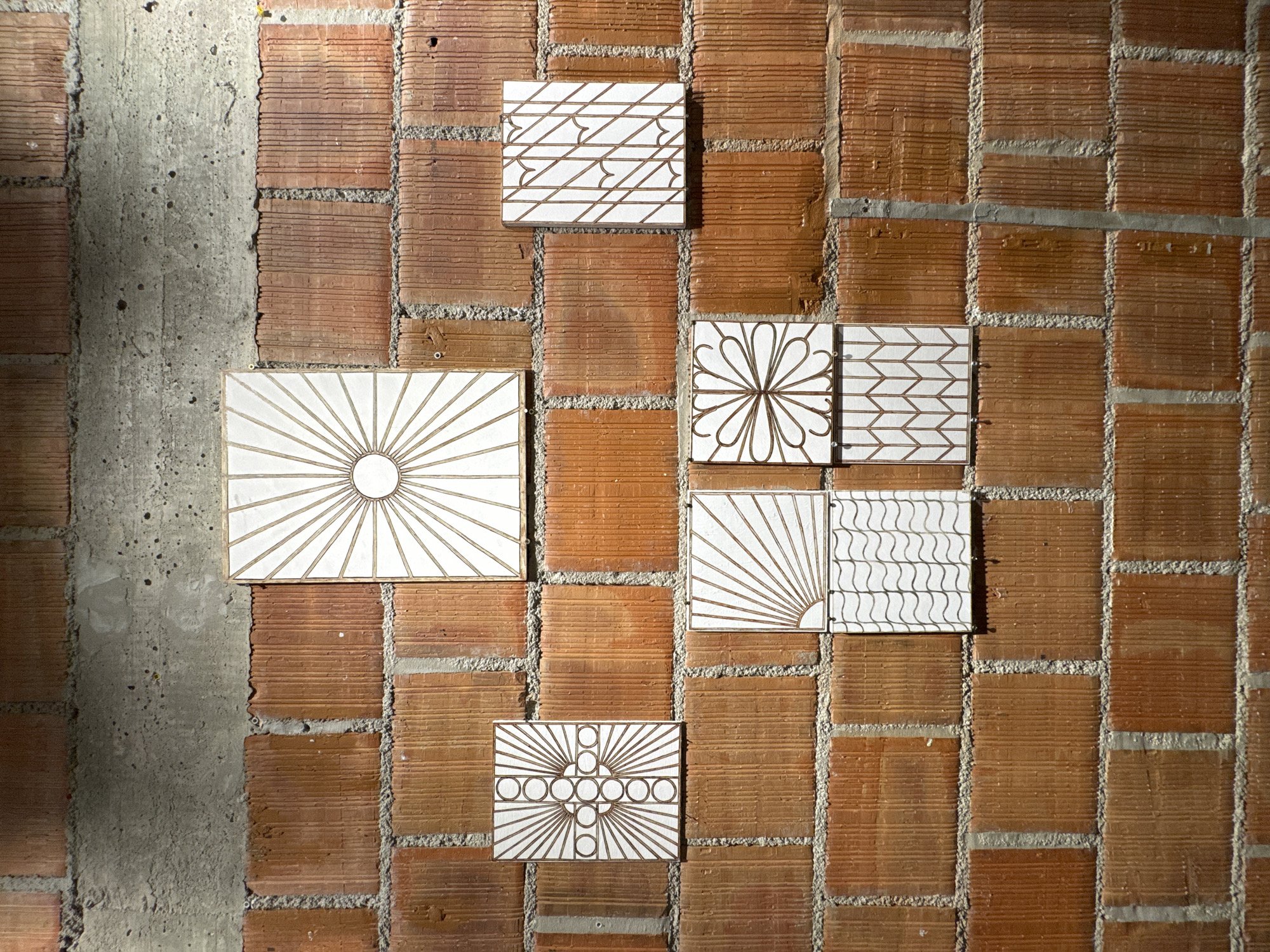 The image shows a brick wall with several decorative tiles attached to it. The wall appears to be a combination of red bricks and a section of gray concrete on the left side. There are five tiles, each with a different geometric pattern in white and brown. The tile patterns include radial designs, line patterns, and floral motifs. The tiles are arranged in an asymmetrical manner on the wall. The lighting in the scene is uneven, with shadows falling across the wall and tiles. The bricks have a grooved texture.
While the image doesn't explicitly provide a location, it's possible the user indicated the city of Budva, Montenegro in their prompt. The architecture could be consistent with similar areas, but that's an assumption.