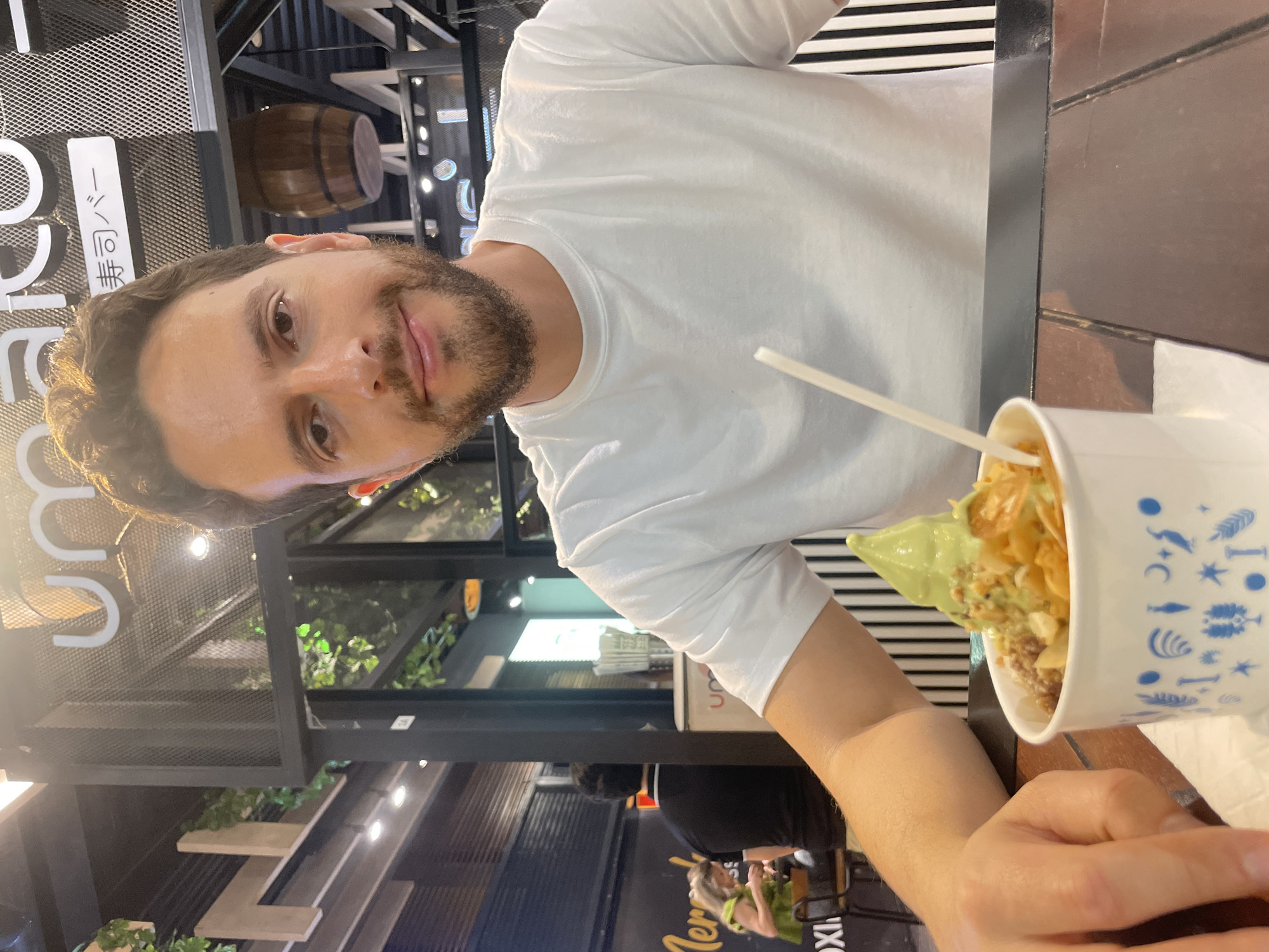 A man with brown hair and a beard is seated at a table, holding a cup of dessert. He is wearing a white t-shirt and appears to be looking at the camera with a slight smile. The cup he is holding contains what looks like soft serve ice cream or yogurt, topped with cereal or nuts. A plastic spoon is sticking out of the dessert.
The man is seated indoors, possibly in a restaurant. The background includes overhead lighting, metal framework, and signs in Japanese. Outside the window, the restaurant "Mercado IX" can be seen. Based on the location of Mercado IX, this image was taken in the city of Puebla, Mexico. Other people can be seen in the background.