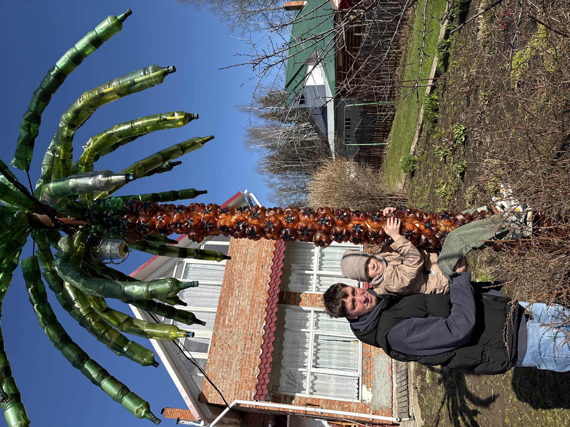 The image shows a man and a child standing near a homemade palm tree constructed from plastic bottles. The man is wearing a black jacket over a blue sweater and blue jeans. He is looking at the camera. The child, dressed in a beige jacket and hat, is being held by the man. They are standing in front of a brick building with white windows and a red tile roof. The palm tree, made from brown and green plastic bottles, is positioned between them and the building. A light fixture is attached to the trunk of the plastic bottle palm tree. In the background, there is another house with a green roof, along with bare trees and a patch of grass. The sky is clear and blue. The ground is covered in dry vegetation and dirt.