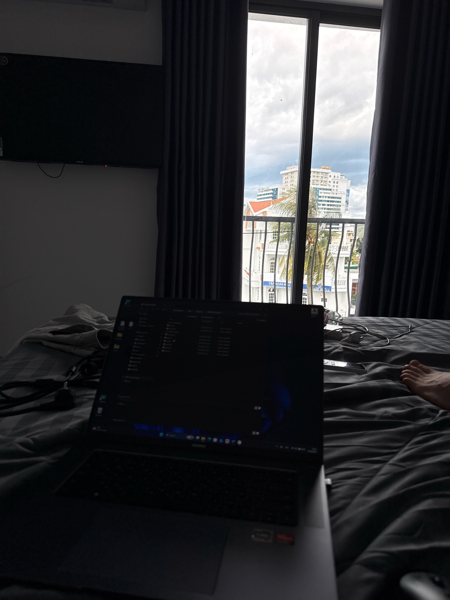 A laptop is open on a bed covered with dark gray bedding, positioned in front of a large window with dark gray curtains. The laptop screen displays a file explorer window. Outside the window, a cloudy sky looms over a cityscape featuring several buildings of various heights and architectural styles, including a prominent multi-story white building with arched windows. Palm trees are visible in the foreground, and a metal railing runs across the exterior of the building visible through the window. A person's foot is visible on the right side of the bed. The scene appears to be set in a hotel room in Nha Trang, Vietnam, given the architectural style and the presence of palm trees. A black flat-screen television is mounted on the wall on the left side of the image.