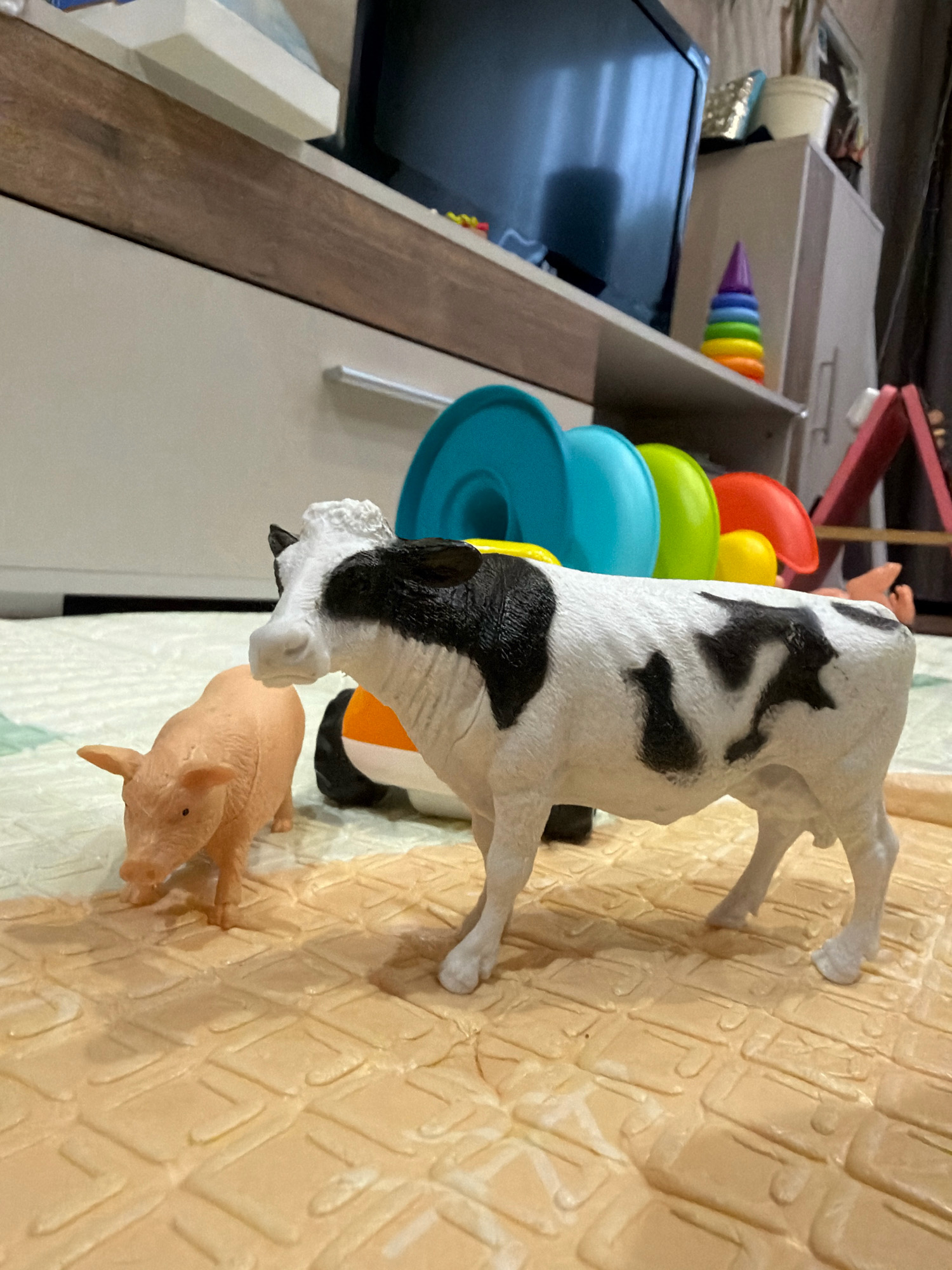 A toy cow and a toy pig stand on a textured mat in front of a white cabinet. The cow is black and white with black spots. The pig is pink. In the background, a television is visible on a shelf above the cabinet, and a stack of colorful stacking toys is on the shelf. The environment appears to be a domestic setting, possibly a child's room or play area.