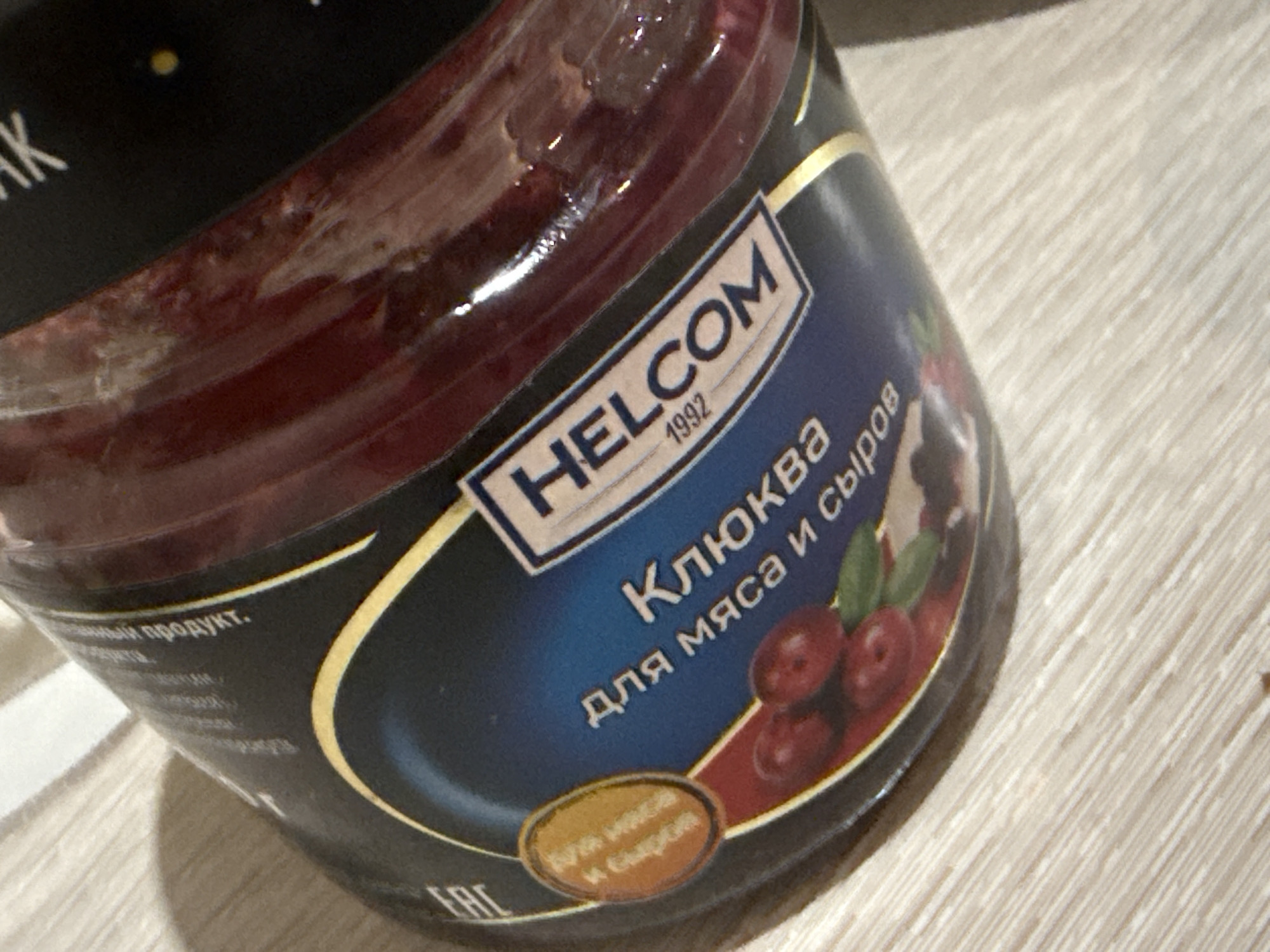 The image shows a close-up of a jar of cranberry sauce, likely "HELCOM" brand, as indicated by text on the label. The jar contains a dark red, viscous substance. The label is predominantly blue and black with gold accents. Prominent text on the label reads "КЛЮКВА" (Klyukva, meaning cranberry in Russian) and "для мяса и сыров" (for meat and cheeses). There is also an oval-shaped inset with text that is difficult to fully decipher but appears to be related to ingredients or use. The jar is placed on a light-colored, horizontally striped surface, resembling a wooden table or countertop. The lighting suggests an indoor setting, possibly with natural light creating shadows on the surface and highlighting the glossy texture of the jar. The scene is devoid of any contextual information related to Dubăsari, Moldova.