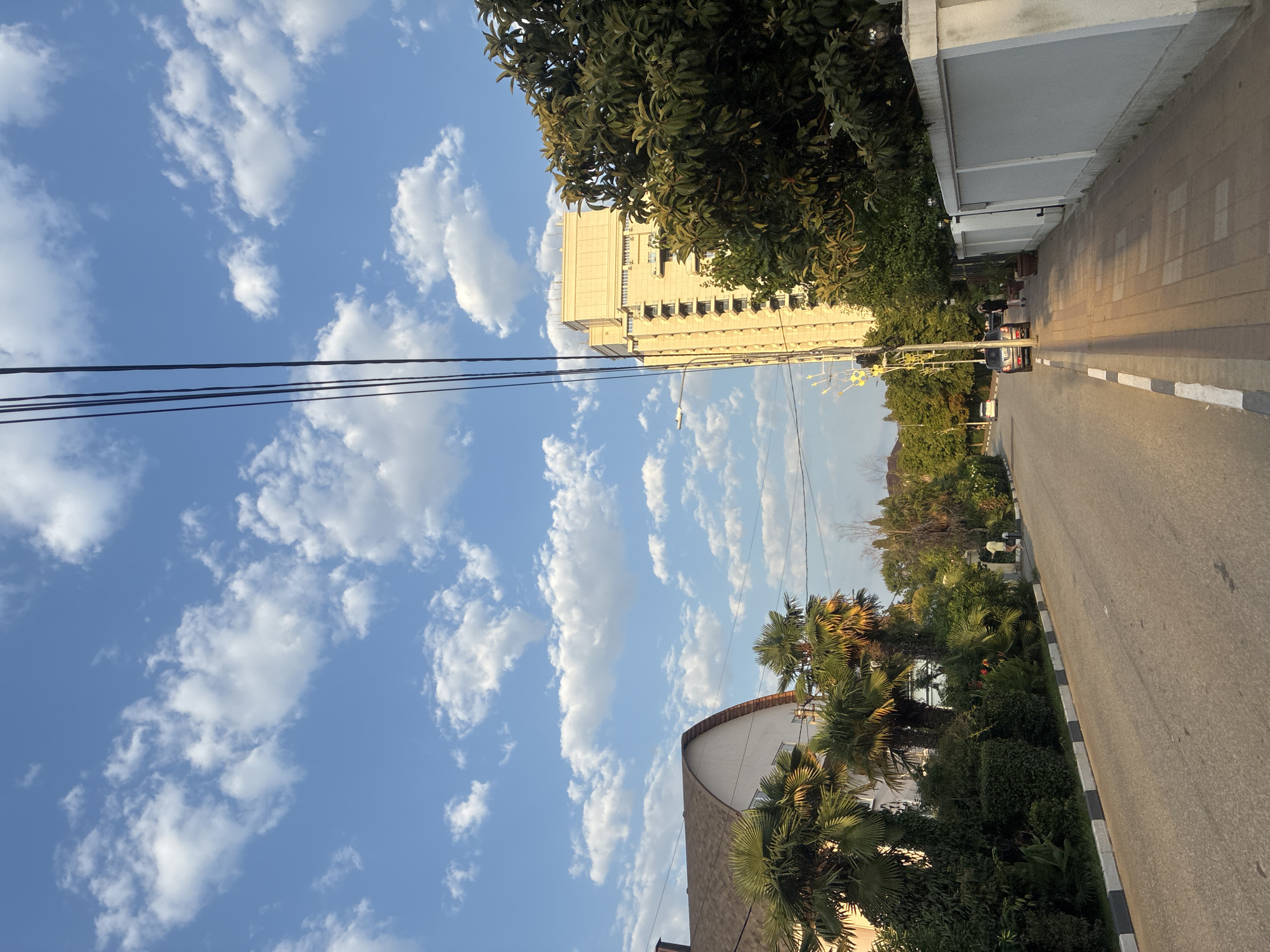 In the city of Sochi, Russia, a paved road stretches through a residential area under a bright blue sky dotted with fluffy white clouds. On the left side of the road, a large, white house with a curved, brown roof is surrounded by lush green palm trees and dense foliage. Across the street, on the right, a tall, light-colored building with many windows stands prominently. In front of the building, a sidewalk paved with interlocking bricks runs alongside a white, stucco-like wall. Several trees with dark green leaves grow in front of the wall. A dark-colored car is parked on the side of the road near the sidewalk, and a person is walking on the sidewalk. Power lines are visible against the sky, running diagonally across the upper portion of the image. The scene appears to be bathed in the warm light of late afternoon or early morning.