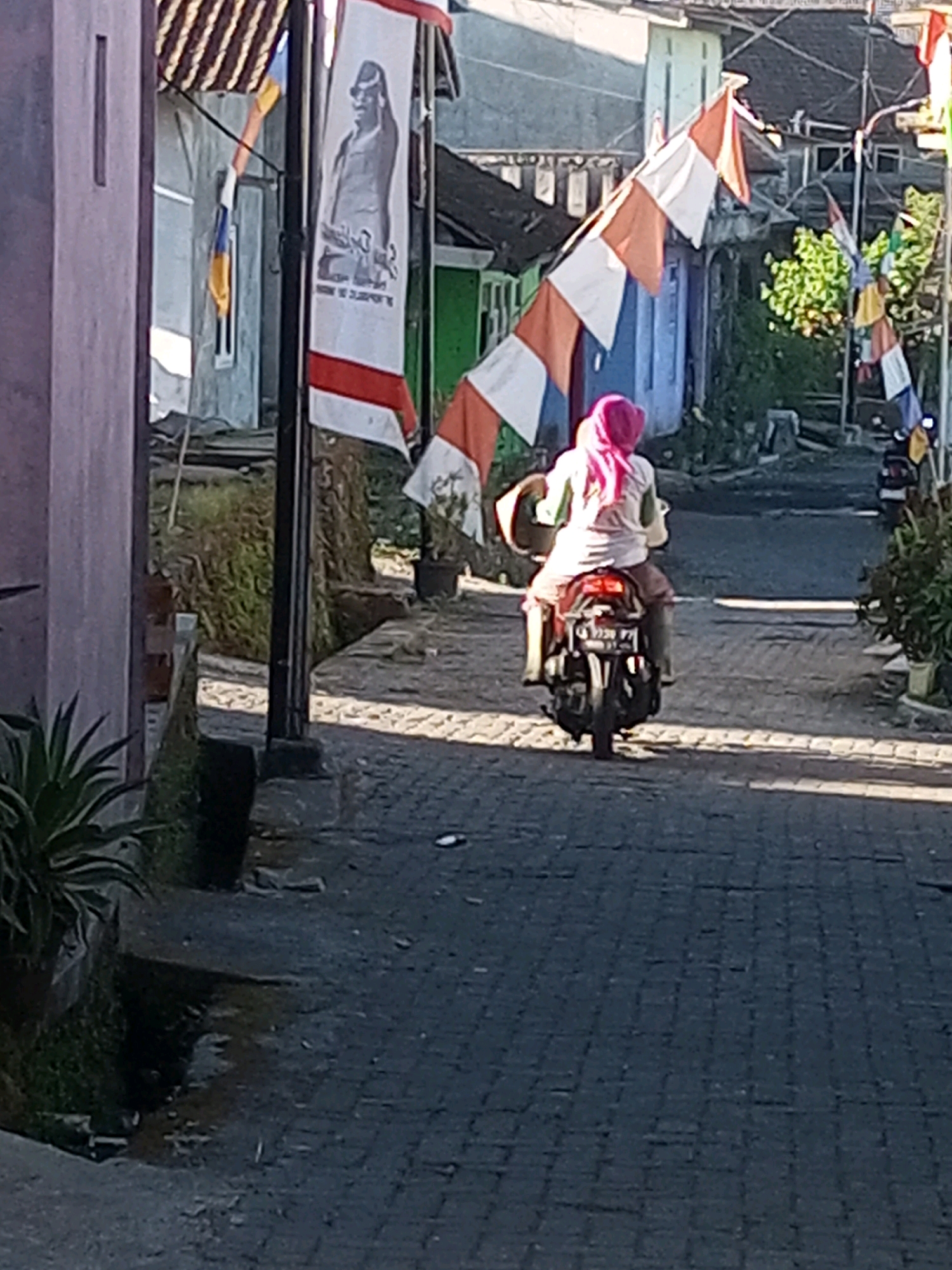 In Andongsili, Indonesia, a person wearing a pink hijab and a white shirt rides a motorcycle down a paved street. The person is carrying a straw hat on their left arm and is seated behind the driver. The motorcycle has a license plate that reads "G 5339 PF". The street is lined with buildings and flags. A tall pole with a banner depicting a person in traditional clothing stands on the left side of the street. Red and white pennant flags are strung across the street. The environment appears to be a village or residential area, with lush greenery visible in the background.