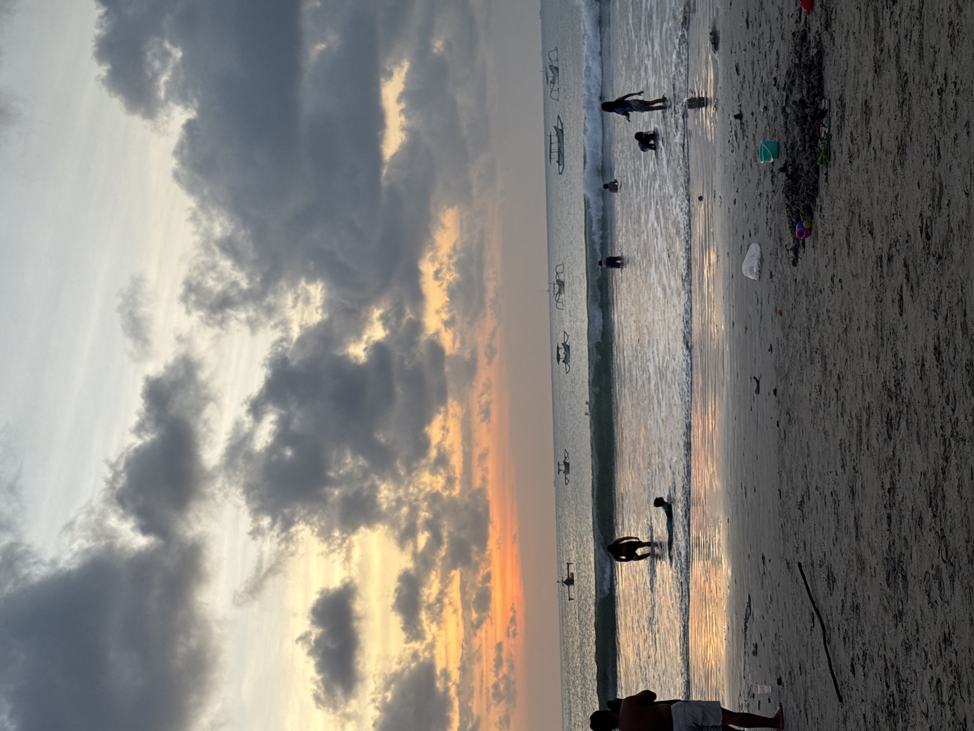 The image depicts a beach scene at dusk with a dramatic sky filled with dark clouds and streaks of orange and yellow sunset hues. Several people are visible on the shore and in the shallow water. Some are wading or playing in the waves, while others are walking along the wet sand. In the distance, several small boats are scattered on the water. The sand on the beach appears somewhat dark and damp, with some debris and scattered objects, including a green bucket, a toy, and a white plastic bag. The overall mood is serene and tranquil, with the soft light of the setting sun reflecting on the water. The location is Kuta Selatan, Indonesia.