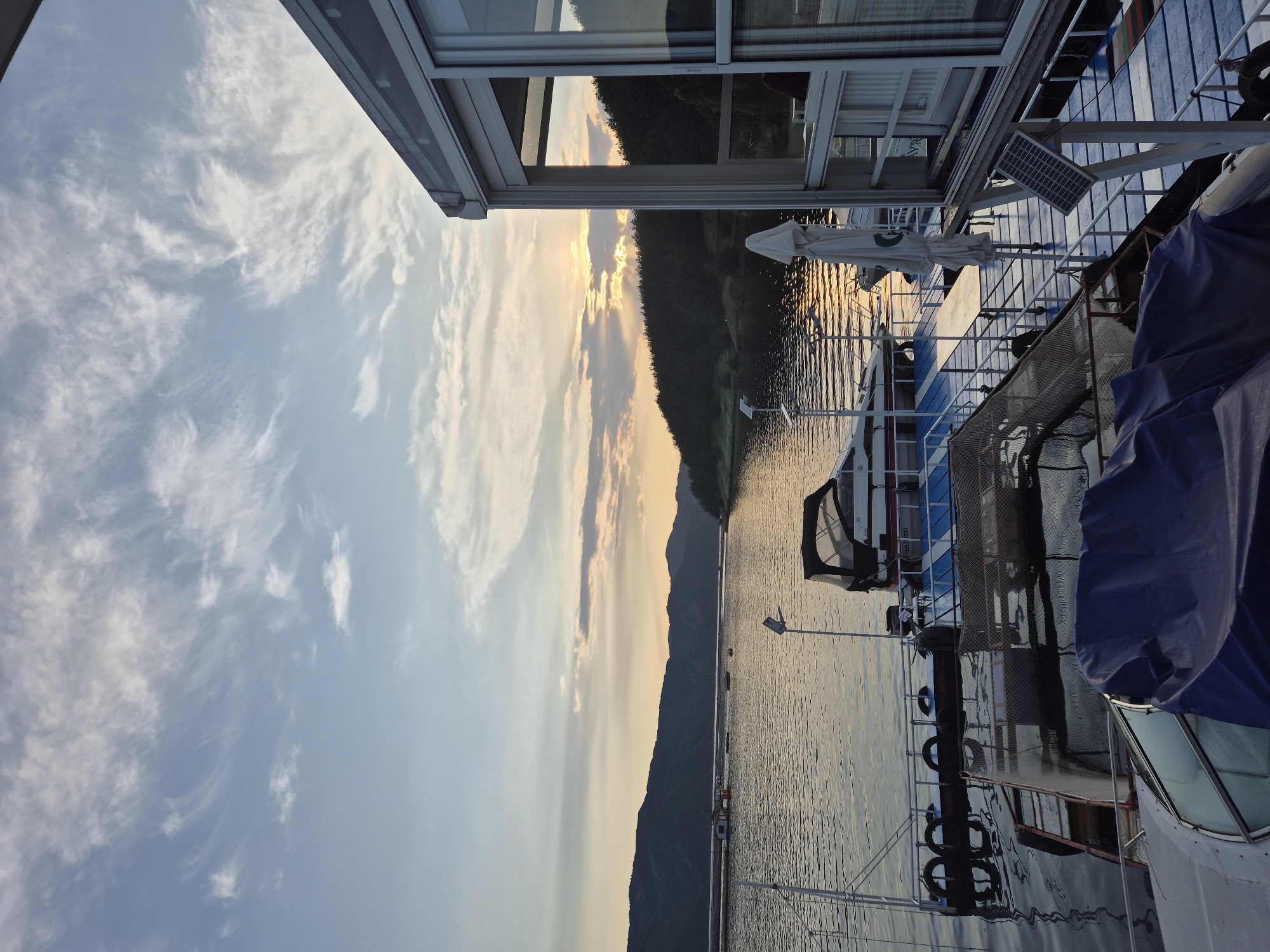 This image captures a serene waterscape at sunset in the city of Brosh, Bulgaria. The low sun is positioned towards the right side of the horizon, casting warm, golden light across the scene, with visible crepuscular rays breaking through scattered cirrus and cumuliform clouds in a light blue sky.
In the background, dark, undulating hills and mountains line the horizon, with a densely forested section visible on the right. A large body of calm water, likely a lake or reservoir, dominates the middle ground, reflecting the warm hues of the sky and the dark silhouettes of the landscape. Several small, indistinct boats are visible on the distant water, along with a small, light-colored floating structure.
In the foreground, a complex of floating docks and structures occupies the lower portion of the image. On the far left, the bow of a boat, partially covered by a blue tarp, is visible. Adjacent to it, a blue and white floating dock with a metal railing extends into the water. Four black tires serve as fenders along its edge. A white motorboat with a black convertible top (a Bayliner, as indicated by faint text) is moored at this dock. A pole with a light fixture extends upwards from the dock. Below this dock, a partially submerged structure covered by a net is discernable in the water.
Further right on the dock, a white, folded patio umbrella stands, and a long, light-colored table is positioned on the dock surface. On the far right, a modern, glass-enclosed structure with white frames is prominently featured. This building, likely a restaurant or lounge, is situated directly on or beside the water. Through one of the large glass windows, a person wearing a red garment is faintly visible inside. A small solar panel is mounted on a pole attached to the base of this structure. The overall impression is one of a tranquil evening at a waterside establishment.