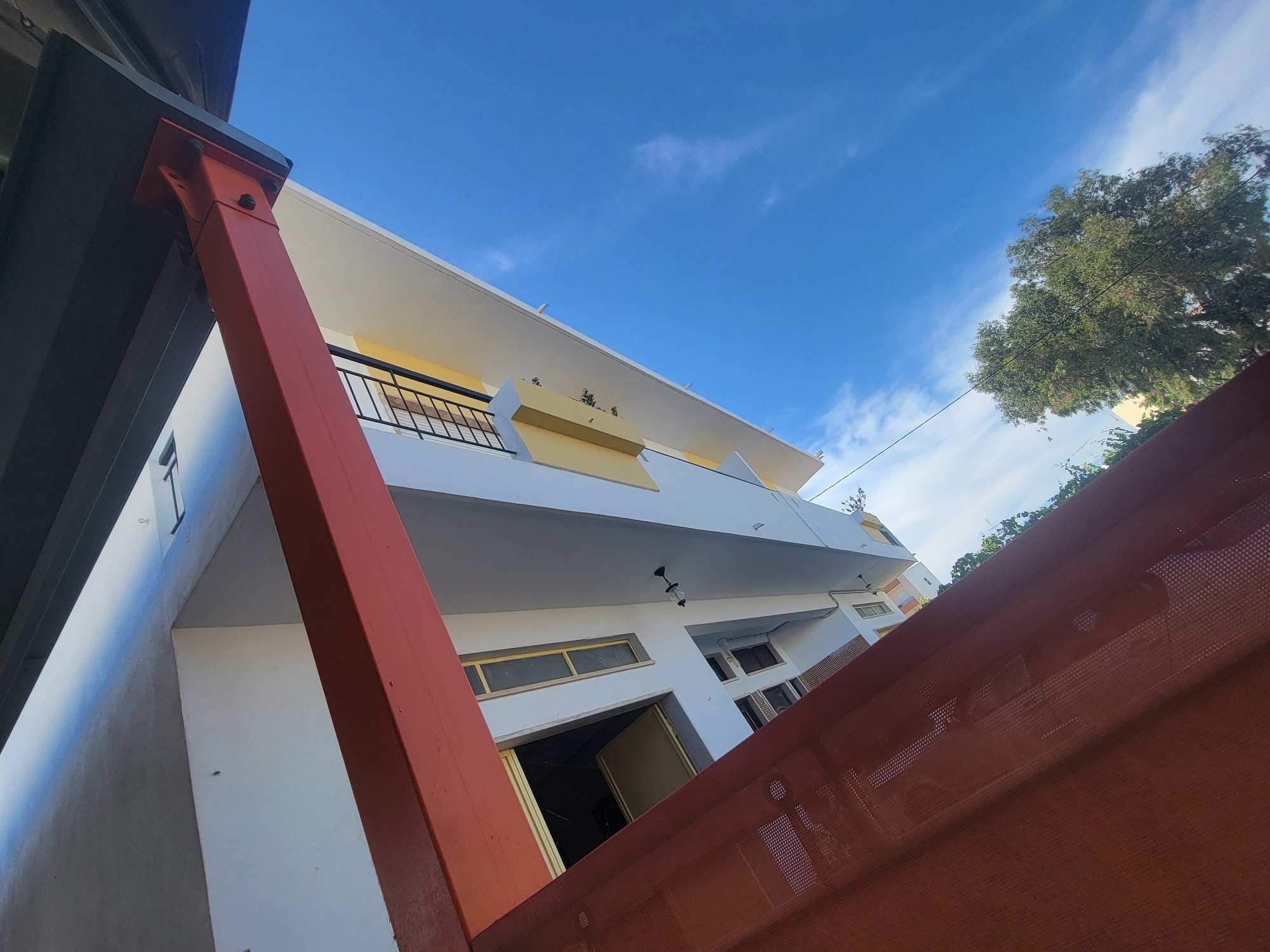 The image depicts a perspective of a modern building with a white facade, a yellow trim, and a dark gray overhang, set against a bright blue sky with scattered white clouds. A prominent bright red structural beam is visible in the foreground, angled diagonally. The building features several windows with yellow frames, and a balcony with a black railing. The architecture suggests a contemporary design. The scene is likely located in Faro, Portugal, given the specific architectural style often found in the region, combined with the clear, bright sky characteristic of the Algarve. The image captures an upward-looking view, emphasizing the building's structure and its relationship with the sky. No people are visible in the frame.