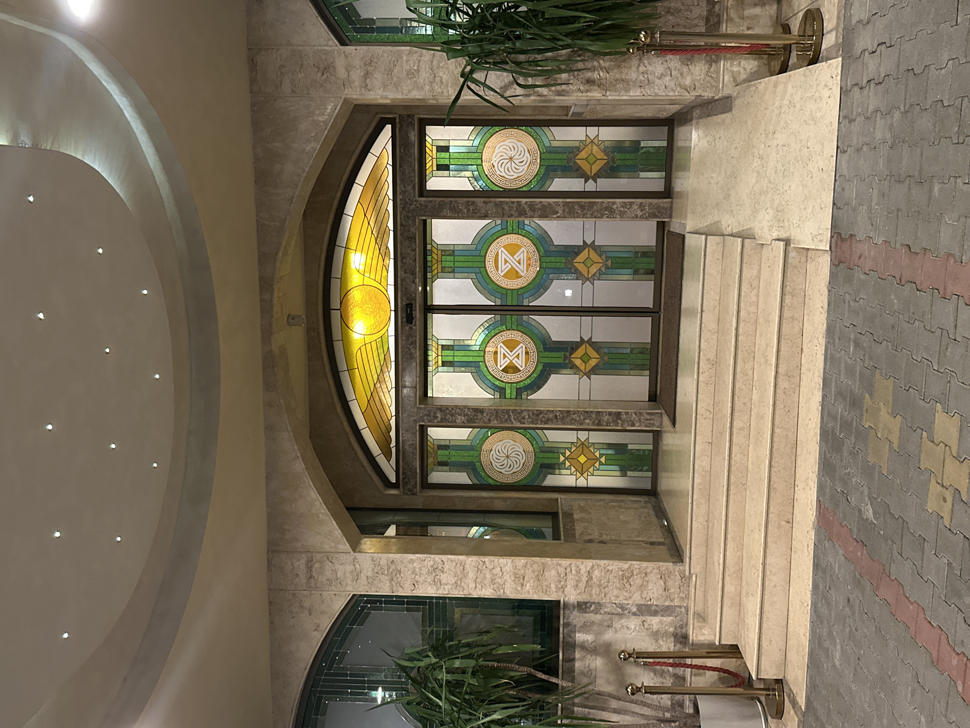 The image displays the illuminated exterior entrance of Hotel Zvezda in Stara Zagora, Bulgaria, at night. The central focus is an arched entryway featuring four glass doors with elaborate stained glass panels. Above the doors, an arched stained glass transom showcases a yellow, sun-like or winged design. The two central door panels each prominently display a circular white 'Z' symbol, while the two outer panels each feature a circular white spiral design, all embedded within a geometric matrix of green, blue, and white glass. Below these circular motifs, diamond-shaped patterns in yellow and green are present. The entrance is flanked by pillars and walls constructed from light-colored, textured stone. Access to the doors is facilitated by a set of three light-colored stone steps on the left and a matching stone wheelchair ramp on the right. Tall potted plants, possibly Dracaena, are positioned on either side of the entrance, each accompanied by a brass stanchion with a red rope. The overhead ceiling is a curved, multi-level structure in neutral tones, fitted with numerous recessed spotlights. The foreground ground surface is paved with interlocking concrete pavers, predominantly grey, with a linear section of red pavers on the left and a diagonal pattern of yellow pavers on the right. No individuals are visible within the scene.