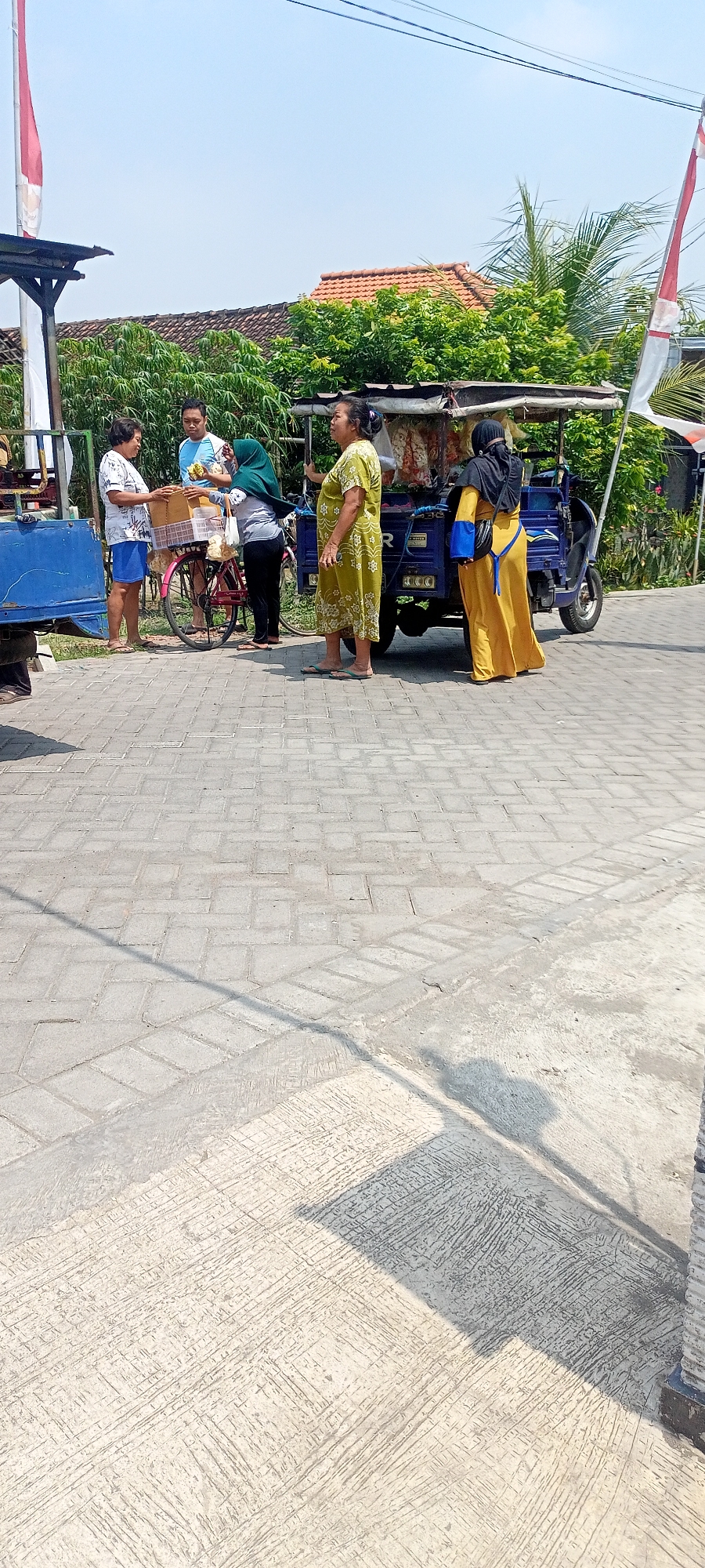 In Gempolklutuk, Indonesia, on a sunny day, several individuals are gathered near a blue motorized vehicle with a canopy, likely a food or goods vendor. A woman in a green patterned dress stands to the right of the vehicle, facing left. To her left, a woman wearing a black hijab and a long yellow dress is seen from the back. In the center, a man in a light blue shirt and a woman in a hijab are interacting. The woman in the hijab is handing something to the man, who is holding a basket. To the far left, a woman in a white t-shirt and blue shorts stands near a blue trailer. A bicycle with a basket is positioned between the man and the woman in the hijab. The ground is paved with light-colored rectangular bricks. Behind the group, lush green trees and a building with a tiled roof are visible. A red and white flag is mounted on a pole to the right. The scene suggests a casual transaction or social interaction outdoors.