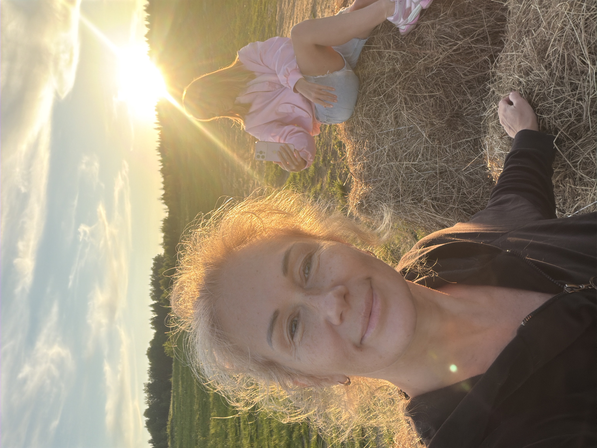 The image captures a warm, golden hour scene in an outdoor setting, likely a field in Керро-2 Снт, Russia. The bright sun, low in the sky, dominates the upper left, casting a strong lens flare and bathing the scene in a soft, golden light. The sky features pale blue patches with scattered clouds.
In the foreground, a woman with light-colored (blonde or light brown) hair is visible, looking towards the camera with a gentle smile. Her hair is backlit by the sun, creating a luminous halo effect. She wears a dark top, possibly a jacket with a zipper.
Behind her and slightly to the left, another female is seated on a bed of dried hay or grass. She has long hair and is dressed in a pink long-sleeved hoodie, light blue denim shorts, and light-colored sneakers with pink accents. She holds a smartphone horizontally in her hands, suggesting she might be taking a photo or video of the view or of the person in the foreground.
The ground is covered in dry, light brown hay or straw, indicating a rural or agricultural environment. A dark treeline is visible in the distant background, separating the field from the sky. The overall mood is serene and relaxed, reflecting a peaceful moment outdoors during sunset or sunrise. No visible text is present in the image.