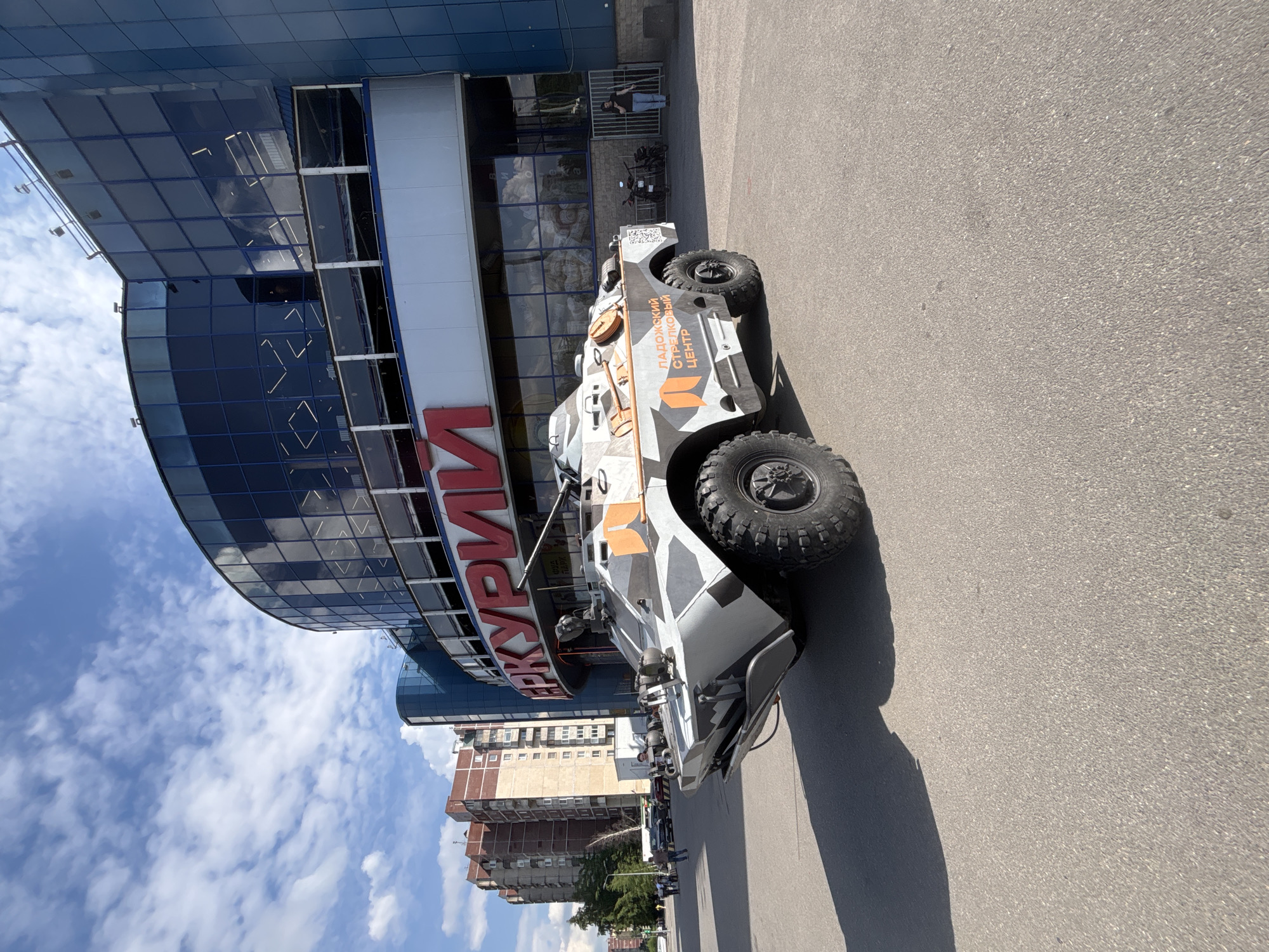 A vertically oriented, sunlit photograph shows a grey and orange camouflage armored vehicle parked on asphalt next to a modern, multi-story building. The sky above is bright blue with scattered white clouds, suggesting a clear day, likely late morning or early afternoon given the distinct shadows cast by the vehicle.
The prominent subject is a large, rugged vehicle, possibly a custom-built armored SUV or a civilianized military-style transport. It features large off-road tires, a heavy-duty front bumper, and a distinctive camouflage pattern of grey, white, and orange, with some black accents. Visible text on its side in Cyrillic reads "ЛАДОЖСКИЙ СТРЕЛКОВЫЙ ЦЕНТР" (Ladoga Shooting Center), accompanied by a logo that resembles a target or stylized letter.
The building adjacent to the vehicle has a curved blue glass facade, reflecting parts of the sky and surroundings. Large red Cyrillic letters are visible on the building's lower section, partially spelling out what appears to be "АРМИЯ" (ARMY). In the background, behind the modern building, several older, multi-story brick and concrete buildings are visible, typical of an urban environment. A person is seen standing near a metal gate or fence behind the armored vehicle. The ground is paved with asphalt. The overall scene suggests a static display or parking arrangement in an urban setting within a Russian-speaking country.