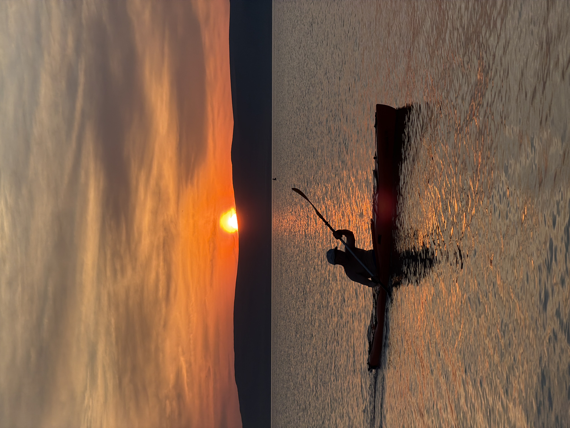 This image captures a serene sunset scene over water. The left half of the image is dominated by a dramatic sky filled with wispy clouds illuminated by the setting sun. The sun itself is a brilliant orange orb peeking over a dark horizon, casting warm hues across the sky. The right half of the image shows a calm body of water reflecting the golden light of the sunset, creating shimmering patterns. In the foreground, a person is visible silhouetted against the water, seated in what appears to be a kayak or canoe. The person is holding a paddle, suggesting they are either about to paddle or are resting. The overall mood is peaceful and tranquil, evoking a sense of quiet contemplation as the day comes to a close. The time of day is clearly sunset due to the intense orange and yellow light and the low position of the sun. There are no visible text elements in the image.
