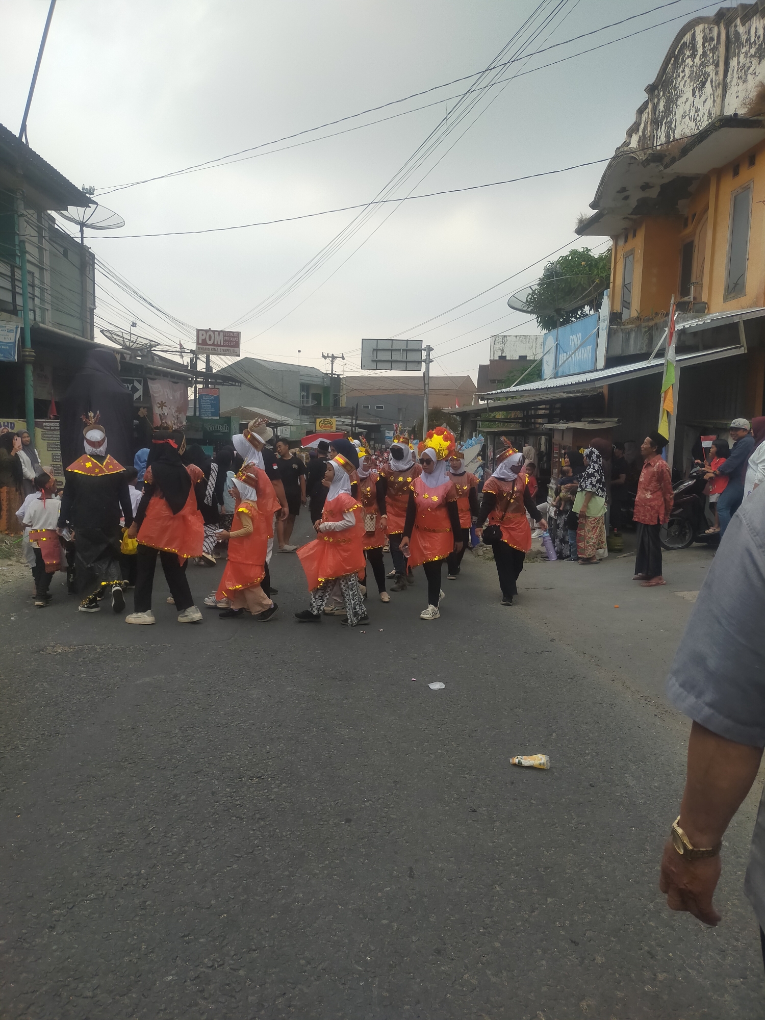 The image depicts a street scene in Tuwel, Indonesia, during what appears to be a parade or festival. A group of people, many dressed in matching orange and black costumes with ornate headdresses, are walking down the paved street. Some individuals are children, while others are adults. The costumes suggest a cultural or ceremonial performance. In the background, there are buildings lining the street, including a yellow building with peeling paint and another structure with a sign that reads "POM". Utility poles and power lines crisscross the sky, which is overcast. People are also observing the procession from the sidewalks. The overall atmosphere suggests a community event taking place in a somewhat urban environment.
