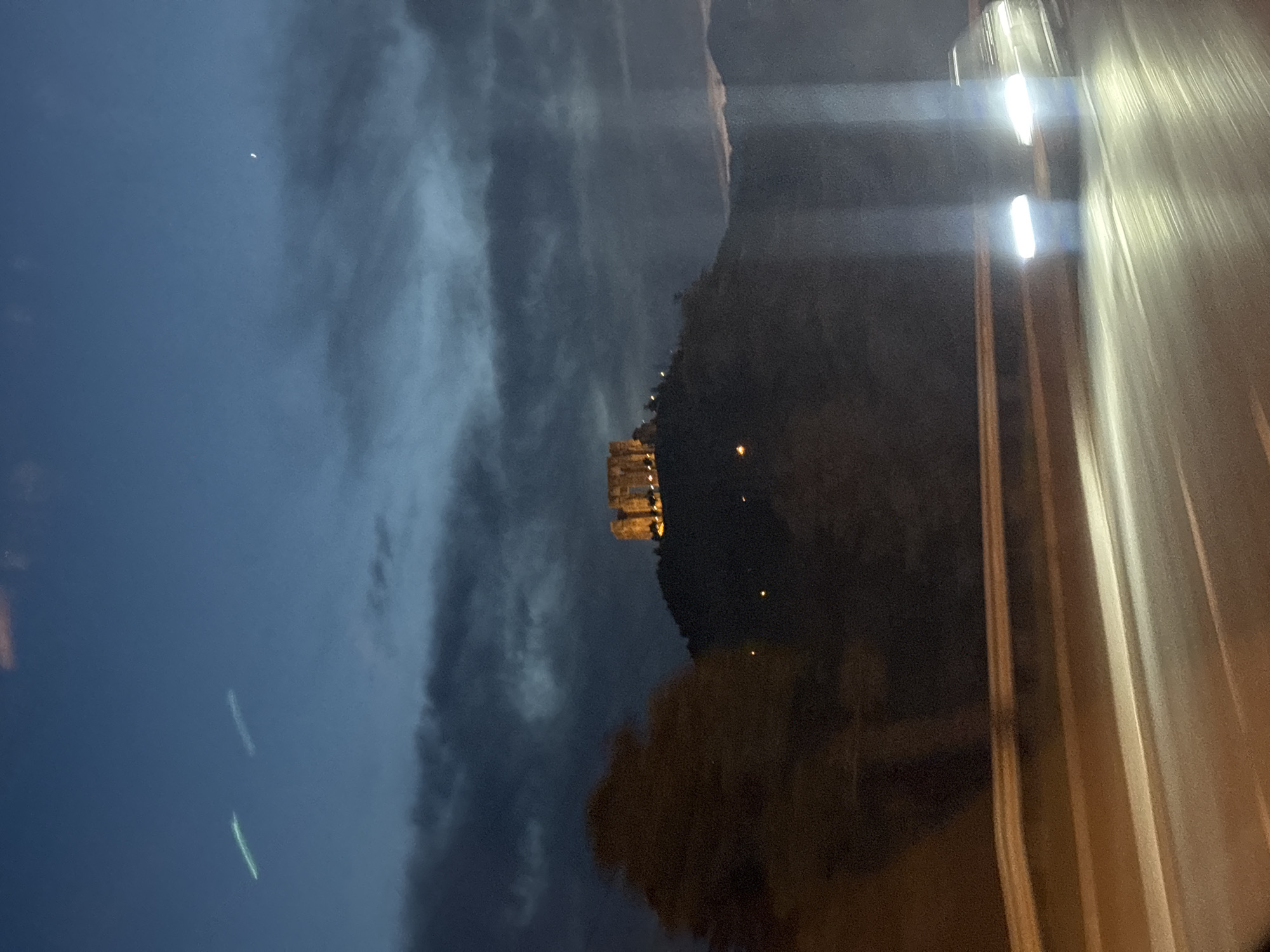 The image depicts a night scene in Tbilisi, Georgia. A portion of the city is visible on a hill, illuminated by warm-toned artificial lights. The sky is predominantly dark with some cloud cover. A few faint celestial objects are visible in the sky. In the foreground, a section of what appears to be a road or highway with vehicle headlights is captured in motion. The image suggests a nighttime view from a moving vehicle, possibly a car or bus.