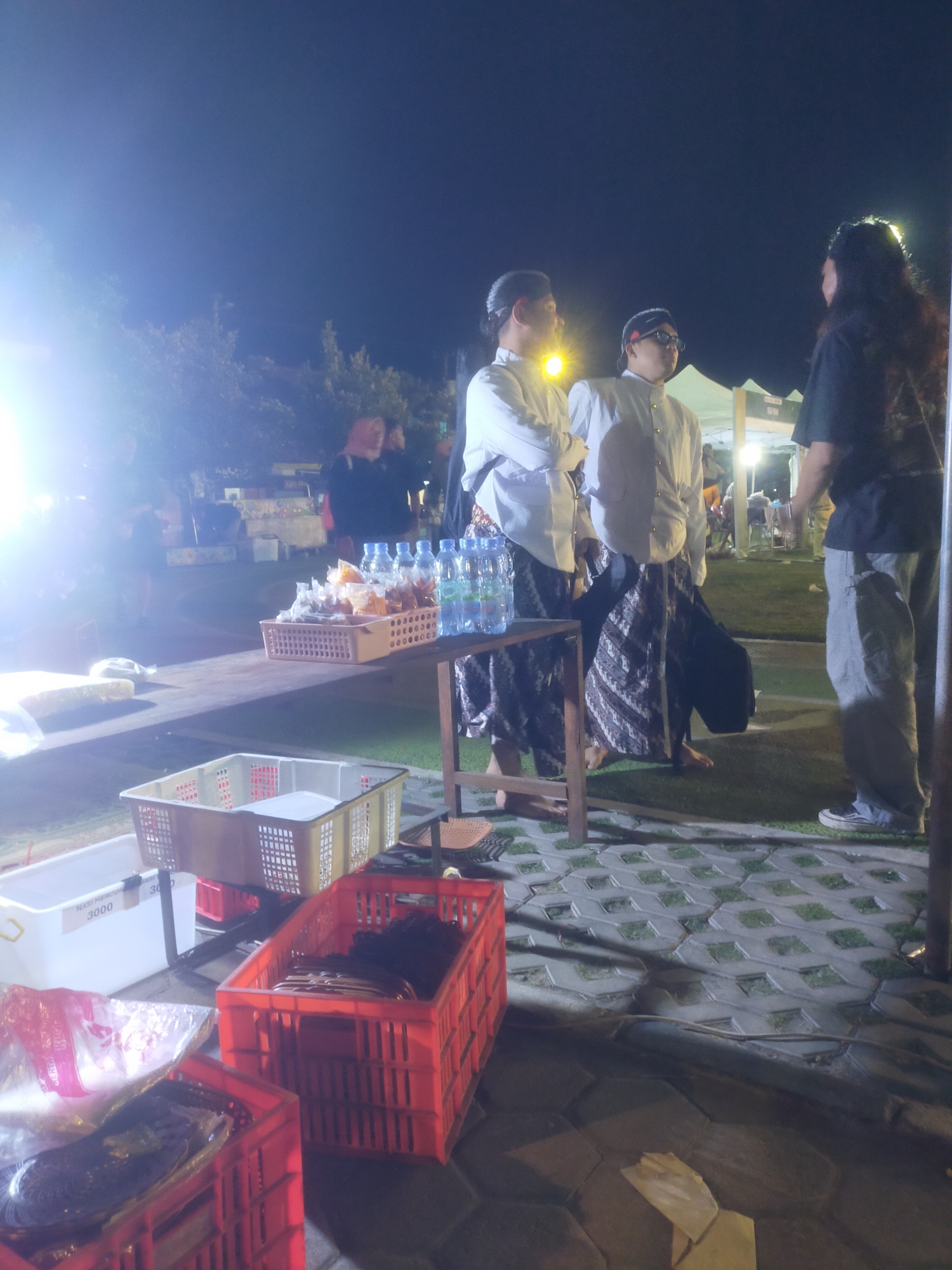 The image depicts an outdoor night scene, likely at a local event or market in Bantul, Indonesia. Three individuals are visible in the foreground, two wearing traditional Indonesian attire consisting of white shirts and patterned sarongs, and the third wearing a dark t-shirt and light grey pants. The two individuals in traditional clothing appear to be conversing, standing near a table laden with bottled water and baskets containing food items. One of the individuals in traditional dress is looking towards the person in the dark t-shirt, who is facing away from the camera.
In the background, there are tents and stalls, suggesting a marketplace or festival setting. Artificial lighting illuminates the scene, creating bright highlights and shadows. In the lower part of the image, several plastic crates are visible, some containing food products. One white crate has "NASI MERCON 3000" written on it, indicating it holds rice with a spicy sauce and costs 3000. The ground is paved with interlocking hexagonal and square concrete blocks interspersed with patches of grass. Trees are visible in the background, illuminated by the artificial lights. The overall atmosphere appears lively and social, typical of a night market or cultural gathering.