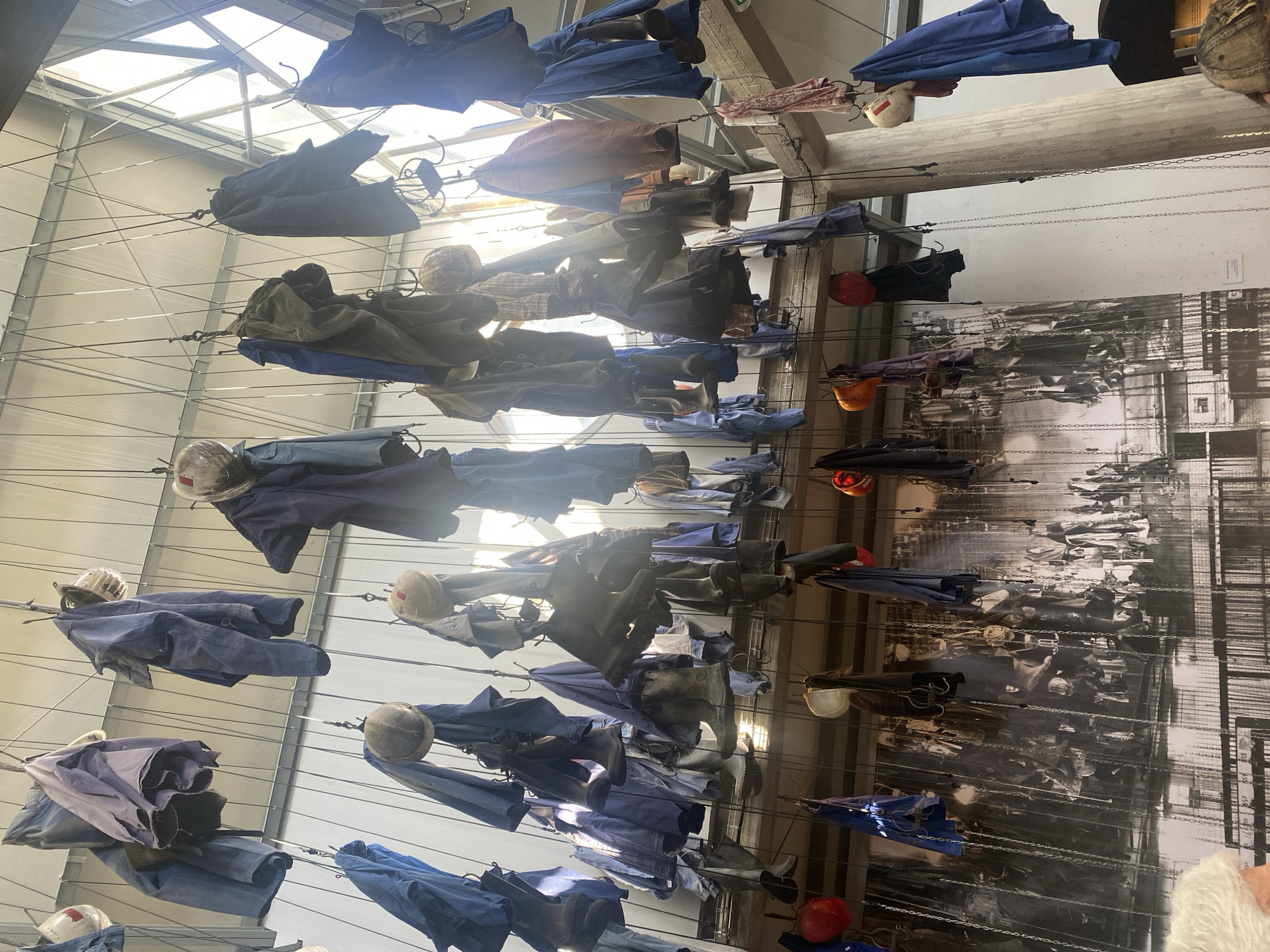 The image captures an art installation at a location in La Motte-d'Aveillans, France. Numerous sets of blue work clothes, including jackets and trousers, are suspended from a complex network of wires. Each set is paired with a hard hat, creating a visual representation of miners or industrial workers. The clothes and helmets are spaced evenly across the frame, hanging from the ceiling. Behind the hanging clothes, a large black and white photograph depicting what appears to be a crowd of people lines a wall. The scene is illuminated by natural light streaming through a windowed ceiling, creating bright patches and shadows throughout the installation.
