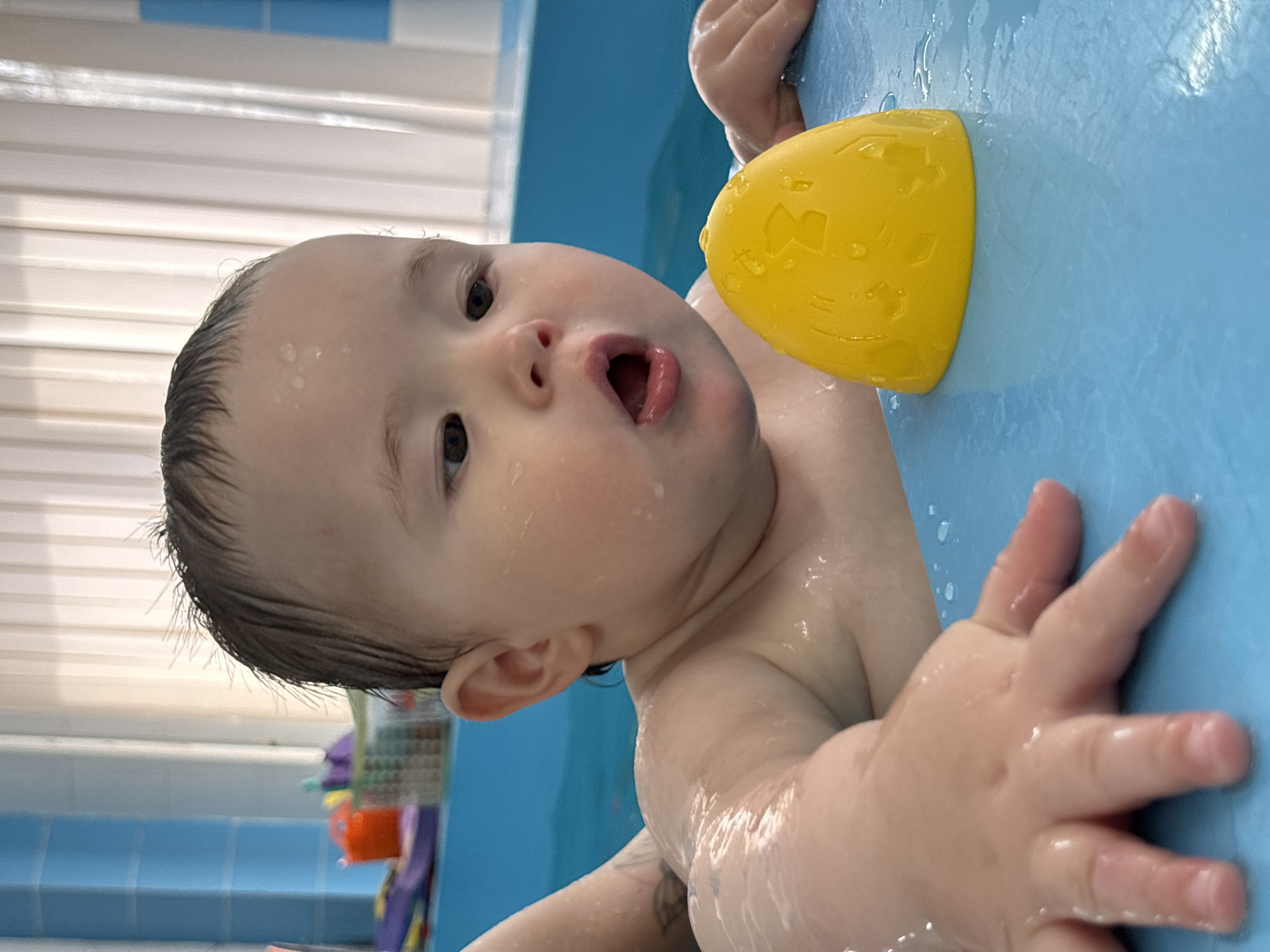 A baby is in a blue tub, grasping the side with one hand. The baby is fair-skinned with wet hair and visible water droplets on their face. The baby's mouth is open. A yellow toy with geometric cutouts is attached to the tub. The background shows a blue tile wall, a window with white blinds, and other baby toys. There's no information in the photo to confirm it was taken in Izhevsk, Russia.