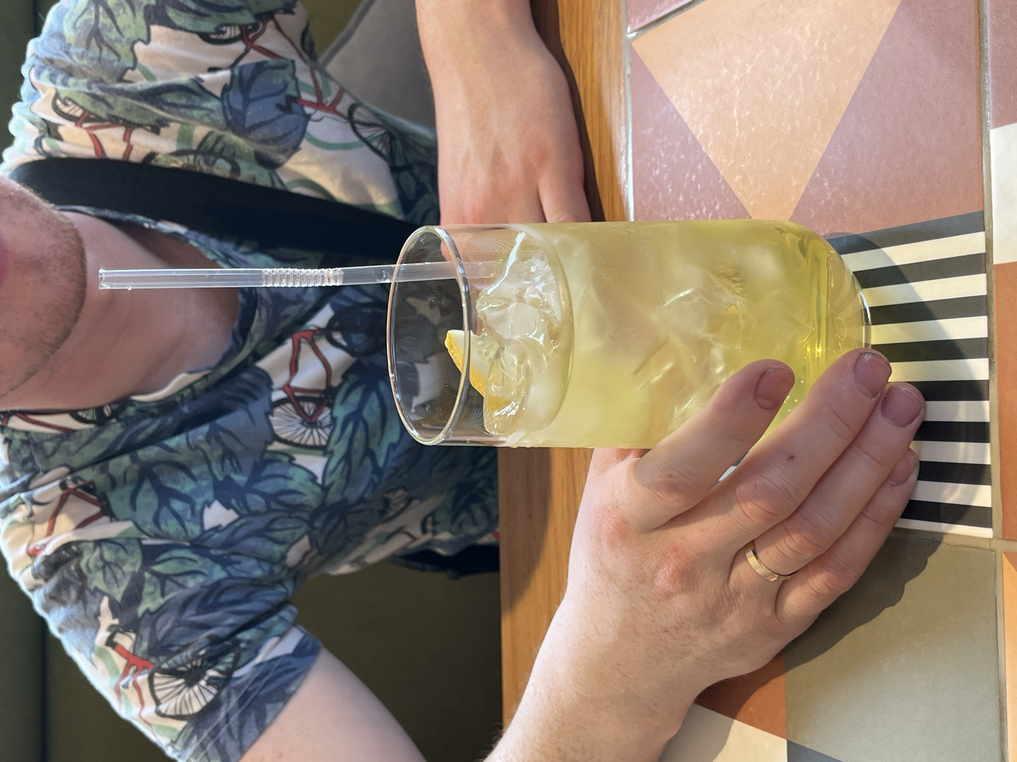 A close-up shows a person holding a glass of a yellow drink with ice and a straw. The person is wearing a patterned shirt with bicycles on it. A gold ring is visible on the person's ring finger. The table has tiles of different colors. The location of the city of Verkhnyaya Pyshma, Russia is not identifiable from the image.