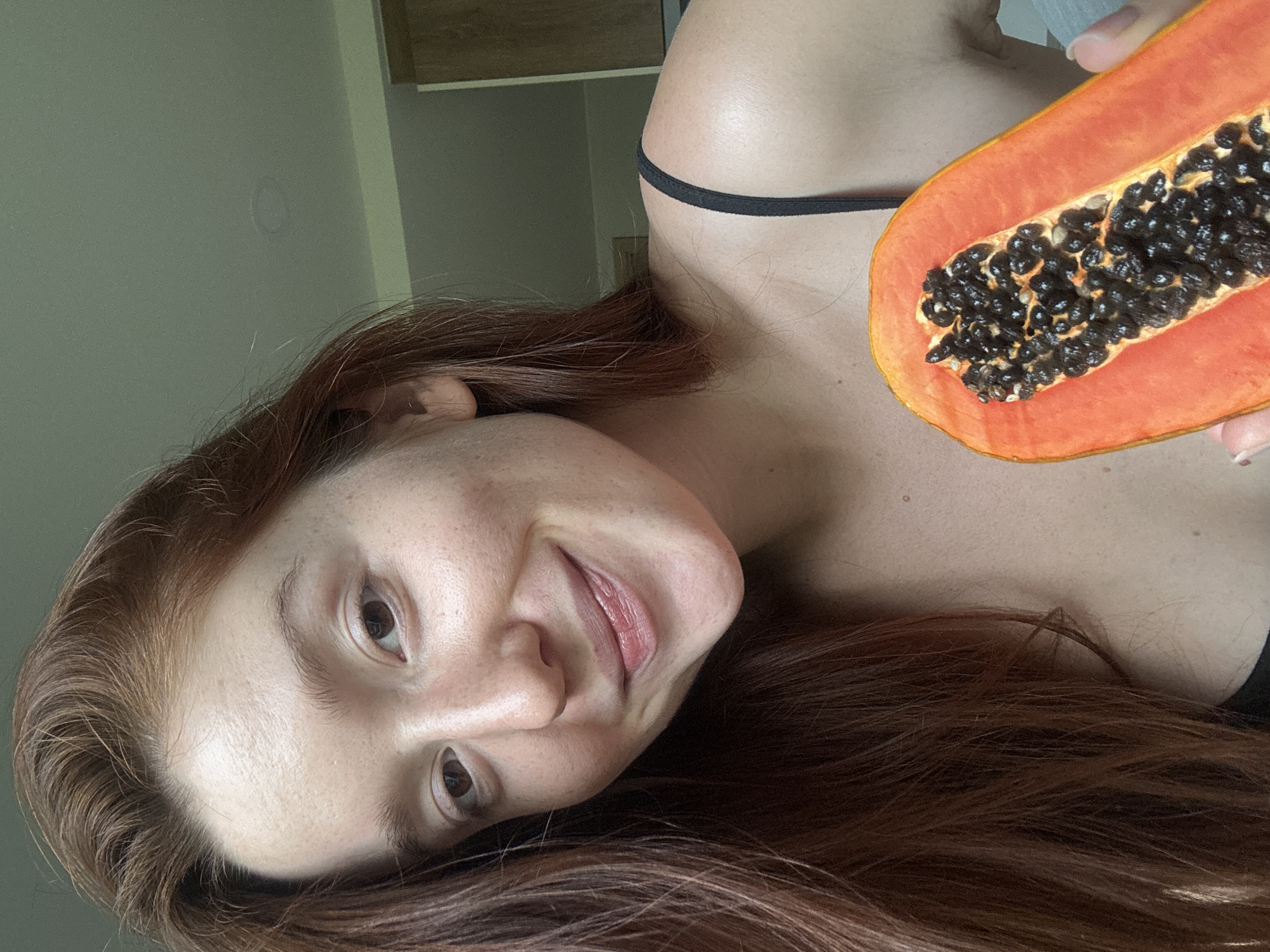 A woman with red hair and freckled skin poses indoors, holding a half-cut papaya filled with black seeds. She is wearing a black tank top. The background shows a light green wall and a partially visible cabinet. The location is assumed to be Choeng Thale, Thailand, based on provided instructions.