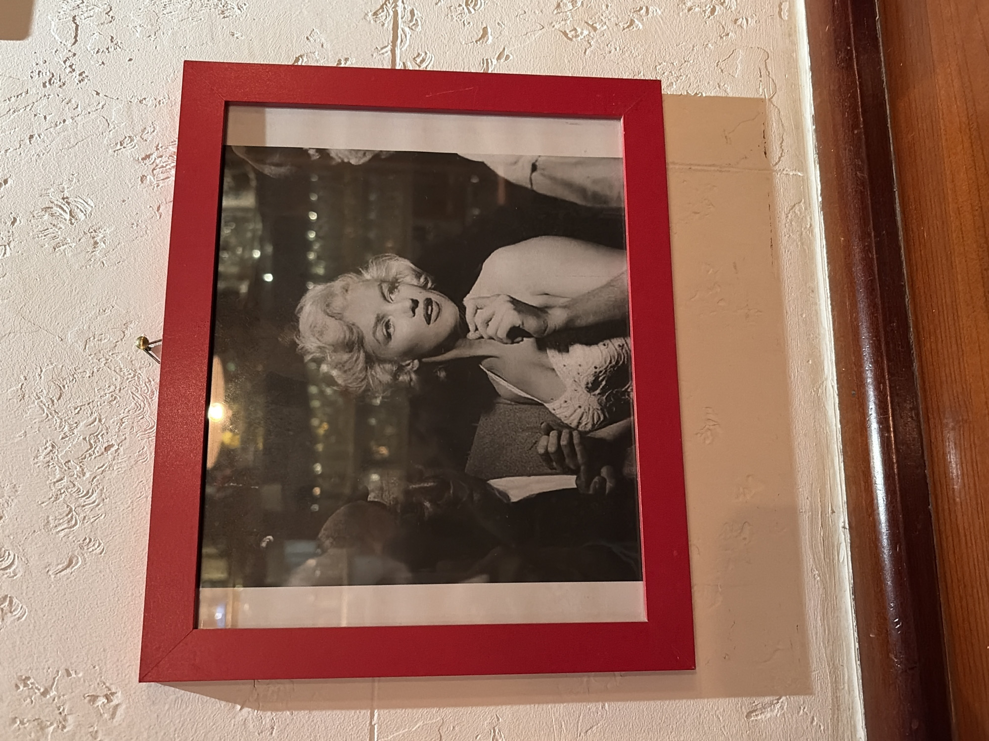 The image depicts a framed black and white photograph of Marilyn Monroe hanging on a textured, off-white wall. The frame is red and appears to be made of wood. The photo features Monroe in a suggestive pose with a man's hands around her neck. The background of the photo appears to be a crowded indoor setting with blurred figures and lights. To the right of the photo is a painted door frame and an adjacent wooden door. The image was taken in the city of Bar, Montenegro.