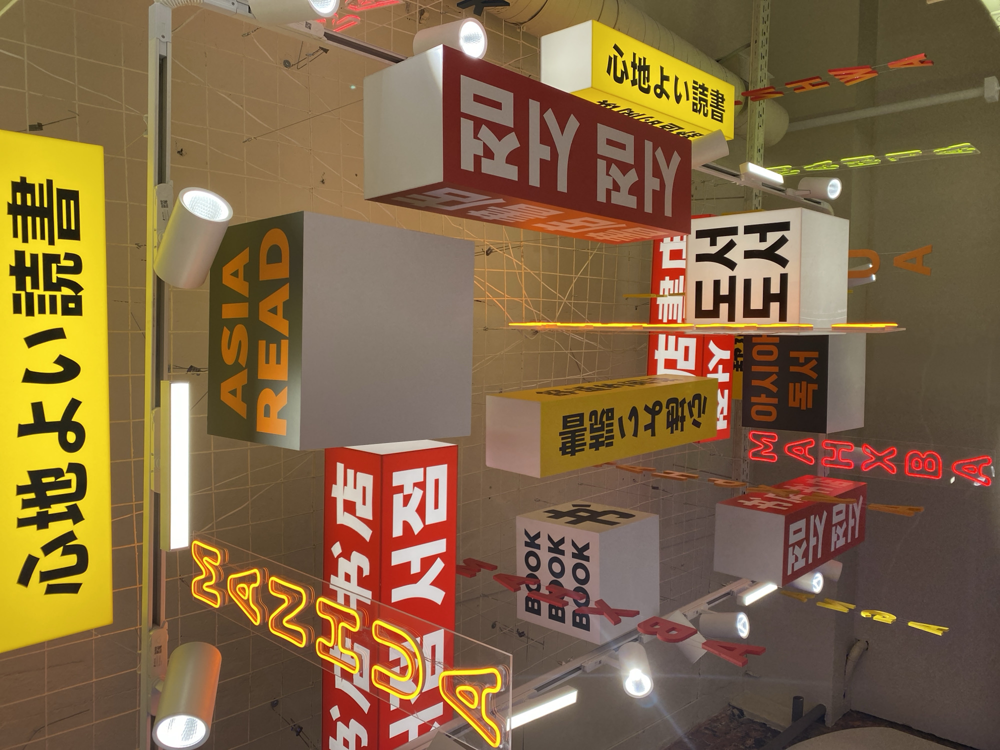 The image depicts a ceiling adorned with numerous hanging signs and lights, suggestive of a retail establishment. Dominant elements include several large, rectangular signs with text in various languages, including Japanese and Korean. Some signs are illuminated with neon, displaying bright yellow and red lettering. One sign at the top reads "心地よい読書" in Japanese, which translates to "comfortable reading." Other signs feature English text like "ASIA READ," "MANGA," and "BOOK BOOK BOOK." Korean text is also visible, such as on a sign that appears to read "만화 서점" (manga bookstore). The overall impression is of a vibrant, visually stimulating environment likely related to books or manga. The specific location in Saint Petersburg, Russia, is not evident from the visual cues within the image itself.