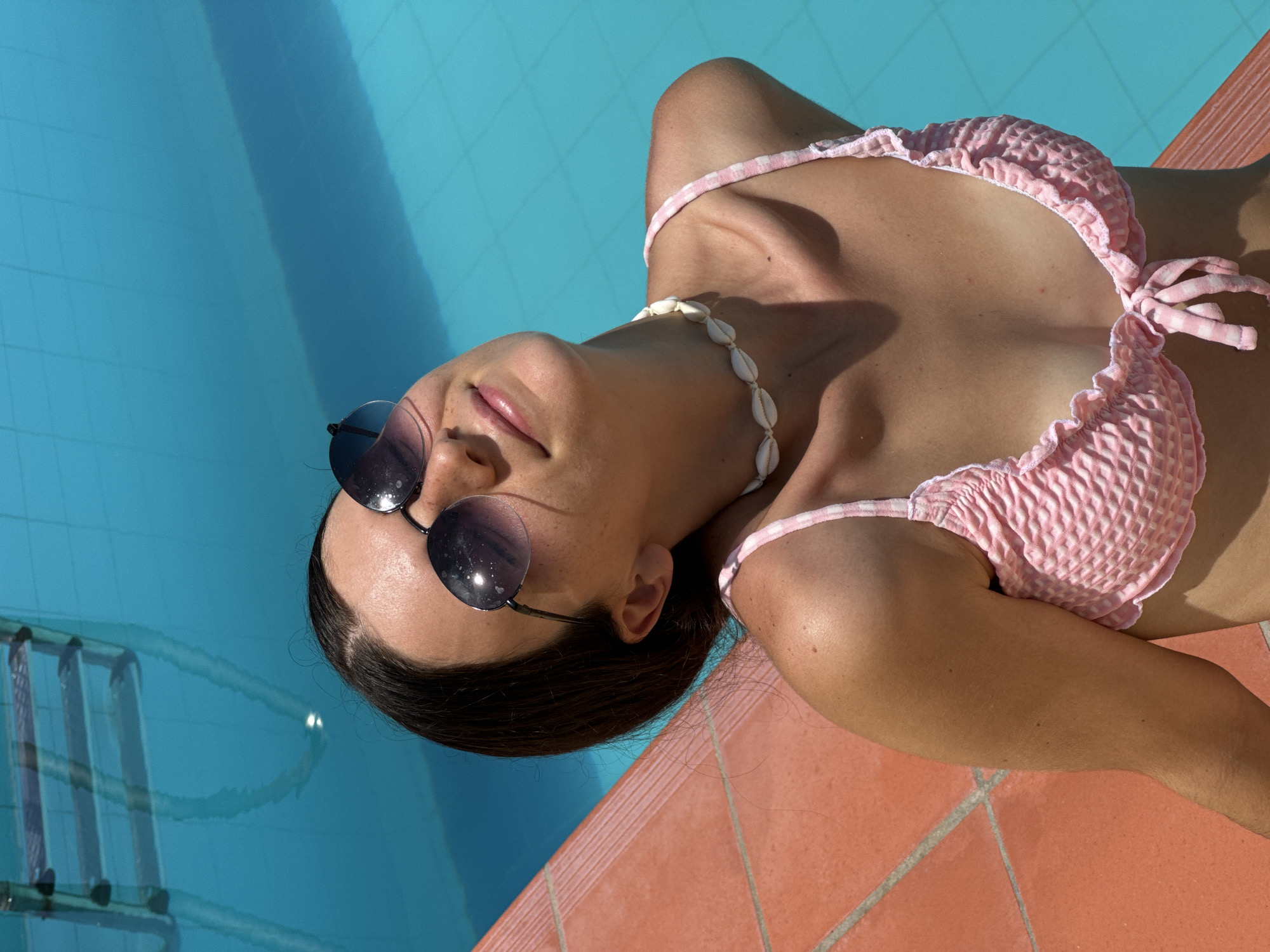 A young woman with dark hair is seated by a swimming pool, wearing sunglasses and a pink and white checkered bikini. She is looking upwards with her head tilted back. She is wearing a seashell necklace. The pool water is bright blue, and the edge of the pool is lined with orange tiles. The scene is lit by bright sunlight, creating shadows on her skin and the water.