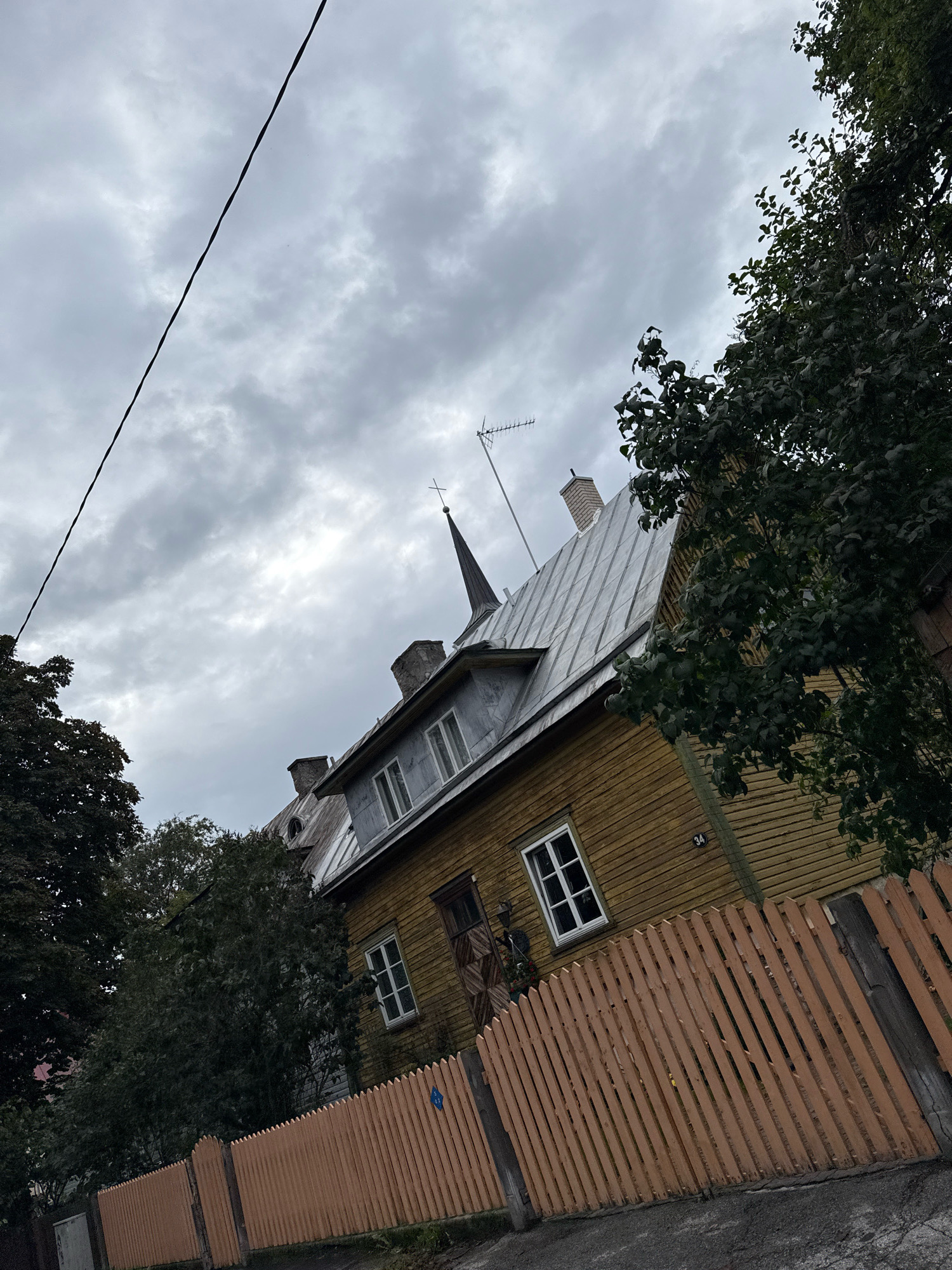 This is an eye-level, medium shot of a wooden house with a light-colored, wooden fence in front of it. The house is painted yellow and has a tin roof. There are several windows on the house, and a door with a patterned design. A small sign with the number "34" is visible on the side of the house. The fence is made of vertical wooden slats and is painted a light orange color. Trees with dark green leaves are visible behind the fence and to the left of the house. The sky is overcast with gray clouds. A black power line is visible in the upper left portion of the image. The scene appears to be in a residential area of Tallinn, Estonia.