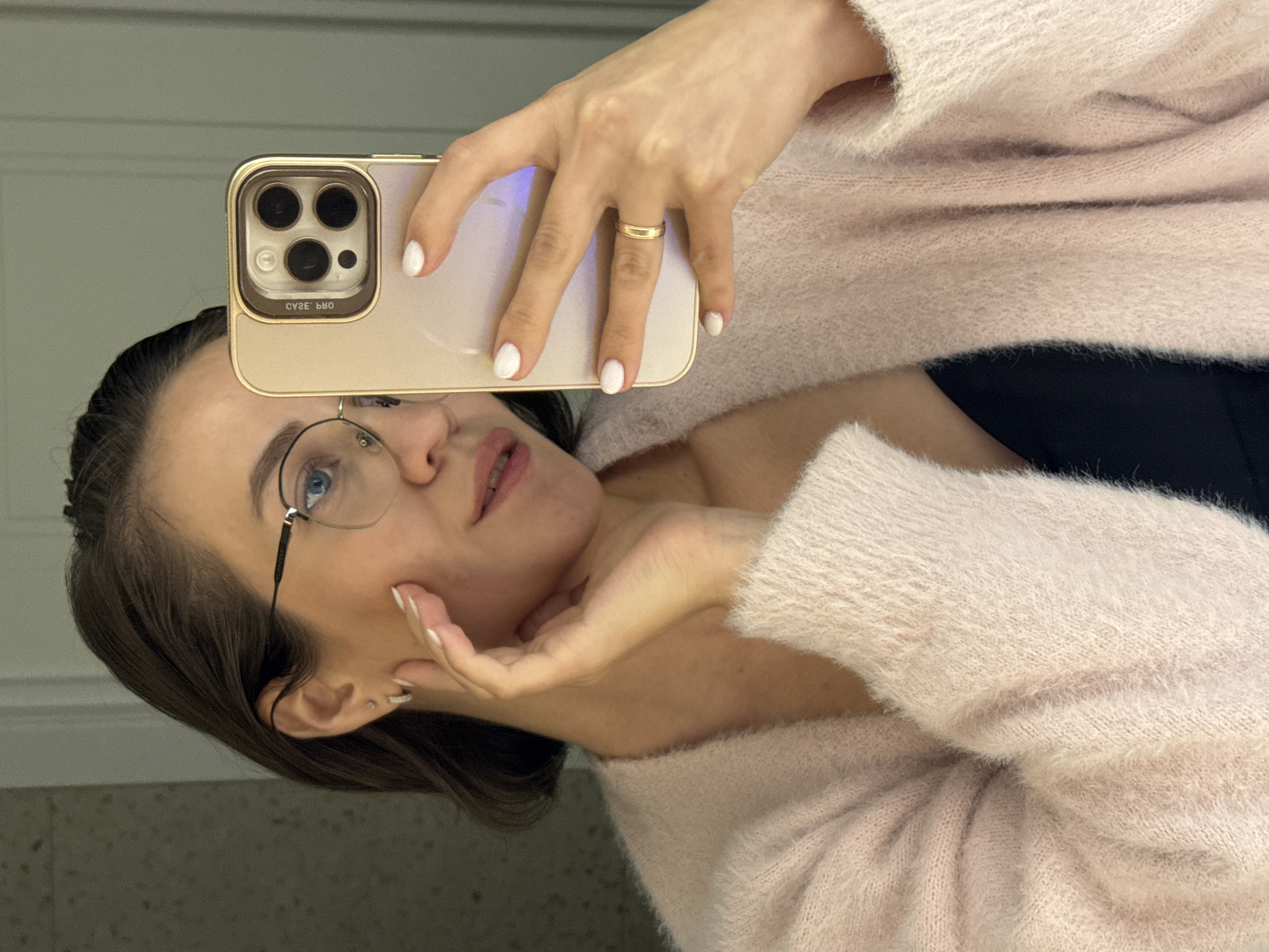 A woman with brown hair, wearing glasses, a pink sweater, and a gold ring, is taking a selfie with a gold iPhone in a room with a light-colored wall. Her left hand is on her face, and she is looking towards the camera. No information is available to determine if the picture was taken in the city of Pervomaysky, Russia.