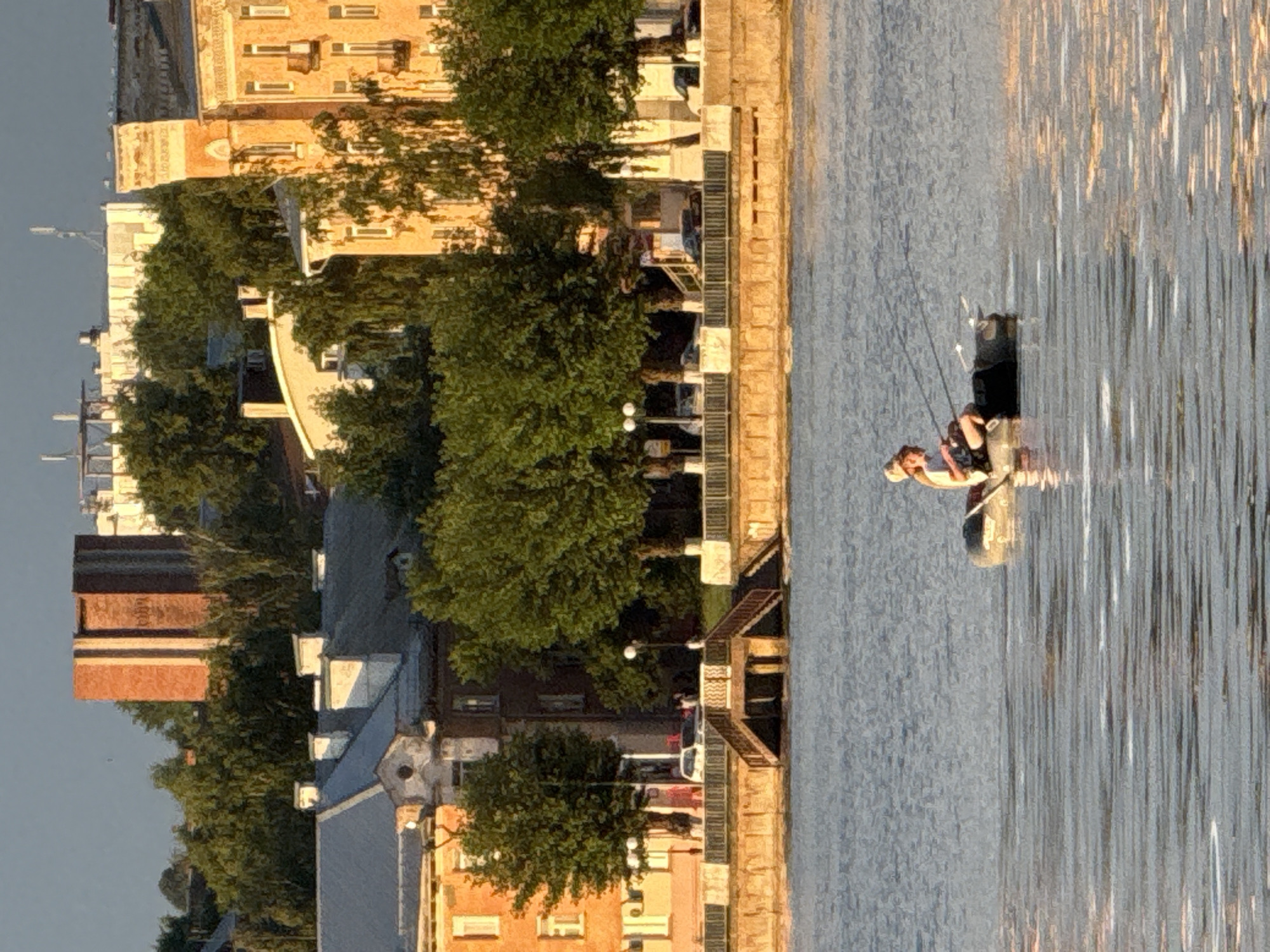 The image shows a body of water, likely a river or lake, in the city of Votkinsk, Russia. In the foreground, a person is fishing from an inflatable boat. The person is wearing a hat and appears to be holding a fishing rod. The water is rippling, reflecting the sunlight. In the background, there are buildings, trees, and a pedestrian promenade. The buildings appear to be old, with a mix of architectural styles. The sky is clear and blue, with some wispy clouds. The overall impression is one of a peaceful, sunny day in a city park or waterfront area.