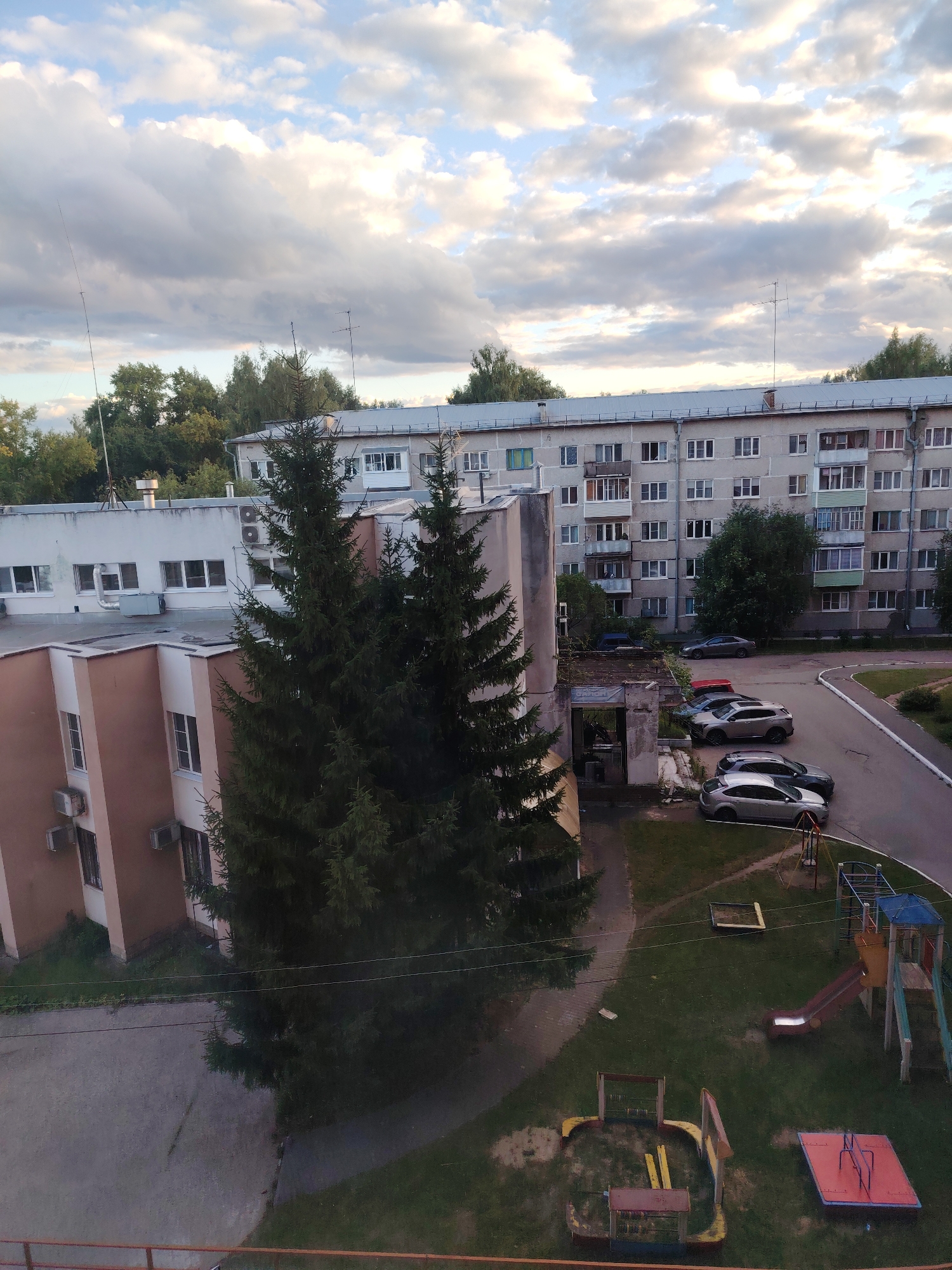 The image shows a view of an apartment building and an adjacent building, possibly a school or office, in the city of Konakovo, Russia. The sky is partly cloudy with dramatic clouds. The apartment building is a multi-story concrete structure with many windows, characteristic of Soviet-era architecture. In front of the apartment building, there is a courtyard with a playground featuring a slide, swings, and a sandbox. Several cars are parked in the courtyard. The adjacent building is lower and has a more modern facade. Two large fir trees are prominently positioned between the buildings. The scene depicts a residential area with typical urban elements.
