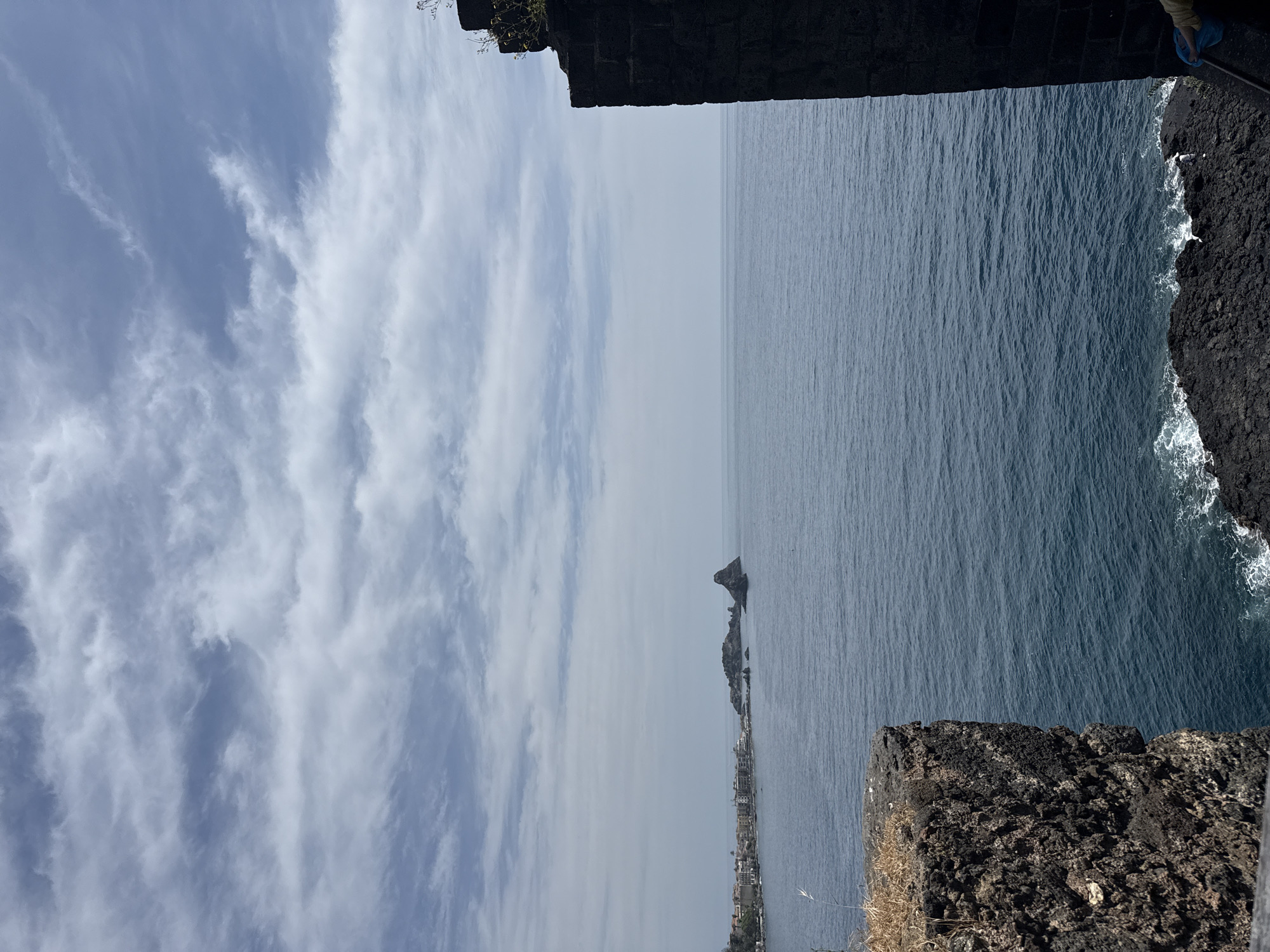 The image depicts a coastal scene. A rock formation is visible in the water near the shore. The sky is partially cloudy. The foreground consists of dark, rocky cliffs. The location appears to be situated in or near a city. The exact city is not discernable.