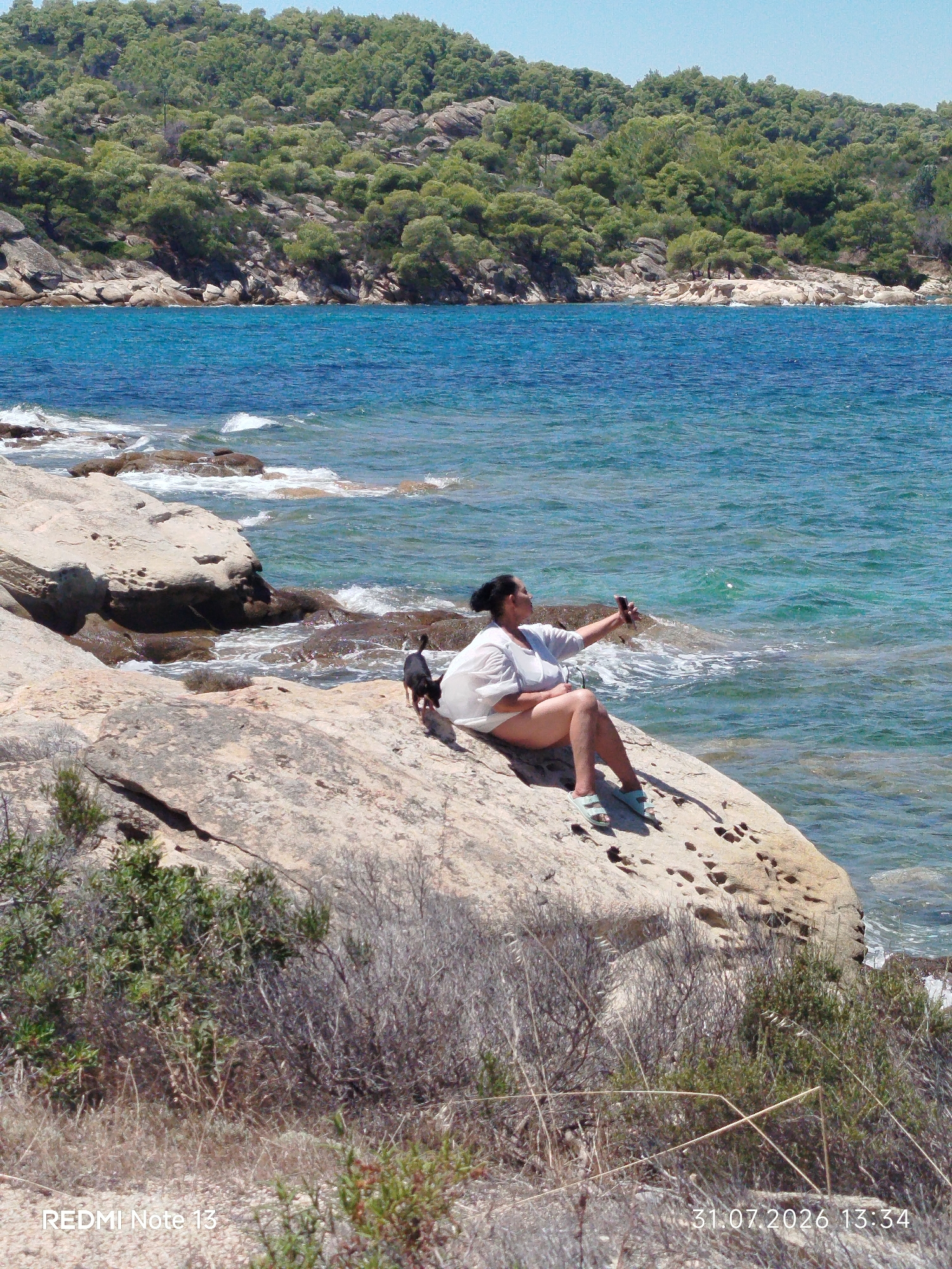 A woman is sitting on a rocky shore in Sithonia, Greece, taking a selfie with her phone. A small black dog is standing next to her, looking towards the water. The woman is wearing a white top and white shorts, and light blue sandals. The sea is a vibrant turquoise, with waves gently lapping against the rocks. Behind the shore, a lush green hillside slopes up towards a clear blue sky. The scene is bathed in bright sunlight, casting shadows on the rocks. The foreground is filled with dry brush and sparse vegetation. The bottom left corner of the image displays "REDMI Note 13", and the bottom right corner displays "31.07.2026 13:34".