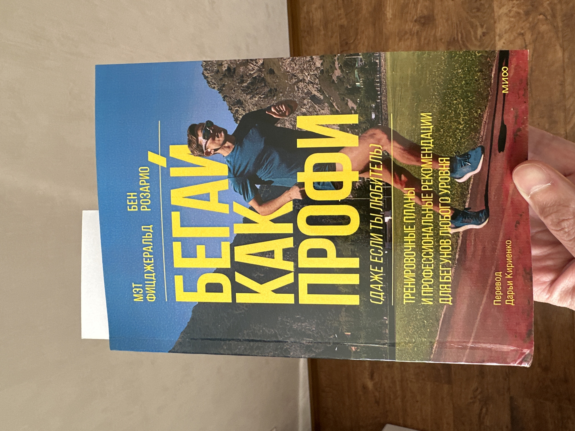 A person is holding a Russian language book titled "Бегай как профи" (Run Like a Pro) in their hand. The cover features a runner in sunglasses against a backdrop of mountains, a track, and a grassy area. The book's authors are listed as Matt Fitzgerald and Ben Rosario, and it promises training plans and professional recommendations for runners of all levels. The text "Даже если ты любитель" (Even if you are an amateur) is also printed on the cover. The translator's name, Darya Kirienko, is included at the bottom right, and the publisher's name, "МИФ", is on the top right. The person holding the book has visible fingerprints. The background is a muted beige or light brown color.